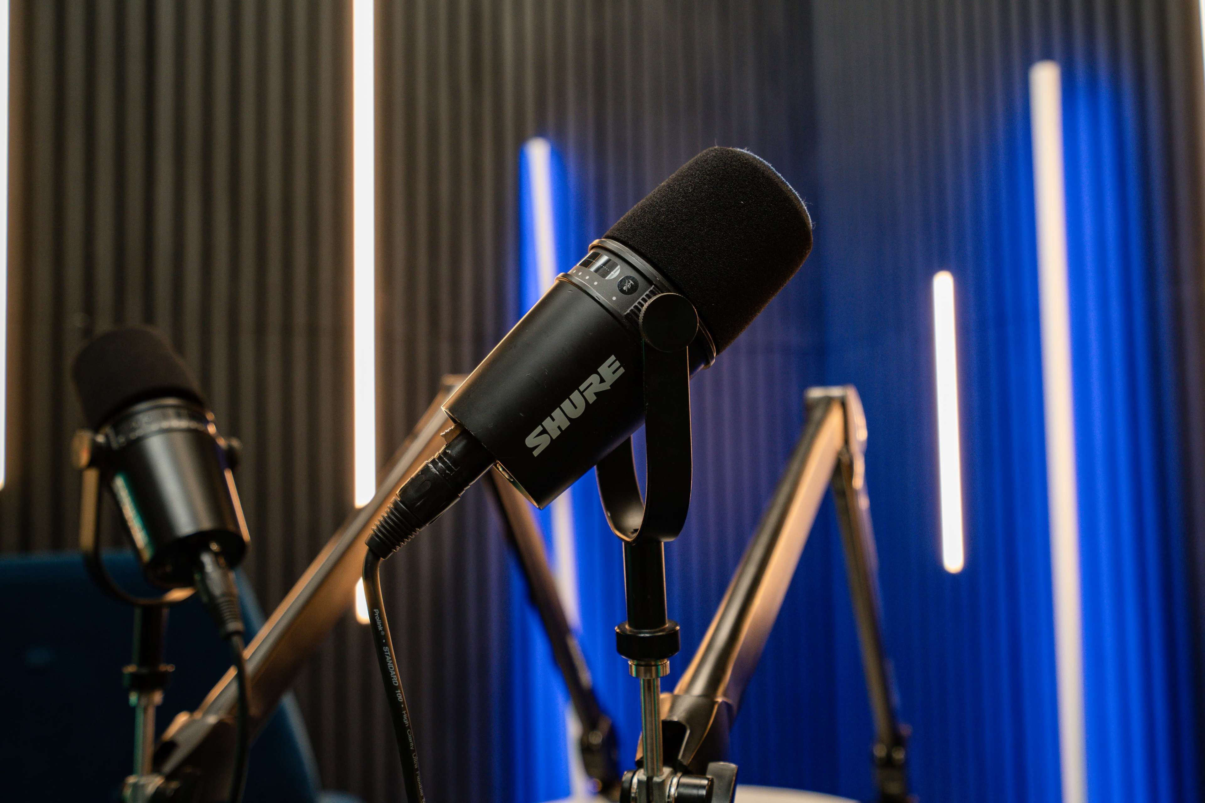 The image shows a close-up of a black Shure microphone mounted on a boom arm in a recording studio with blue and white vertical lights in the background.
