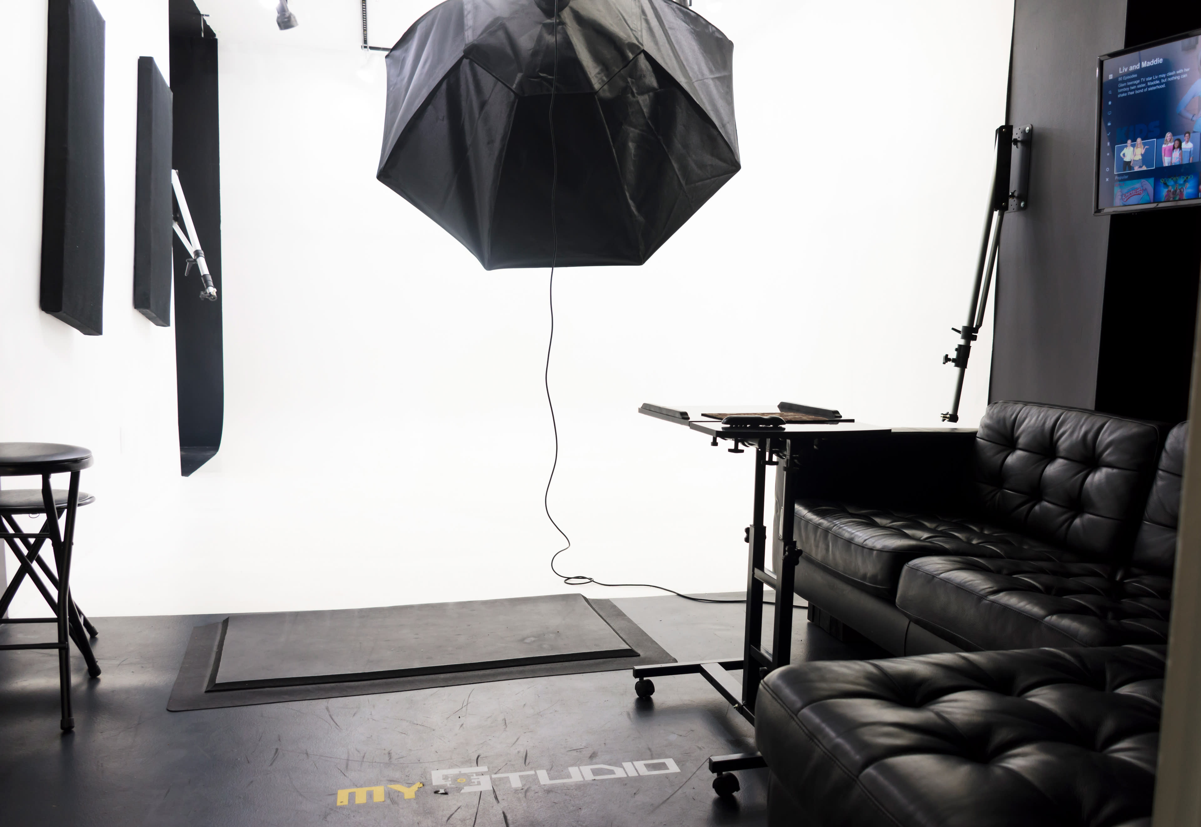 A photography studio with a black umbrella light suspended above a black couch and a table on wheels, against a white backdrop.