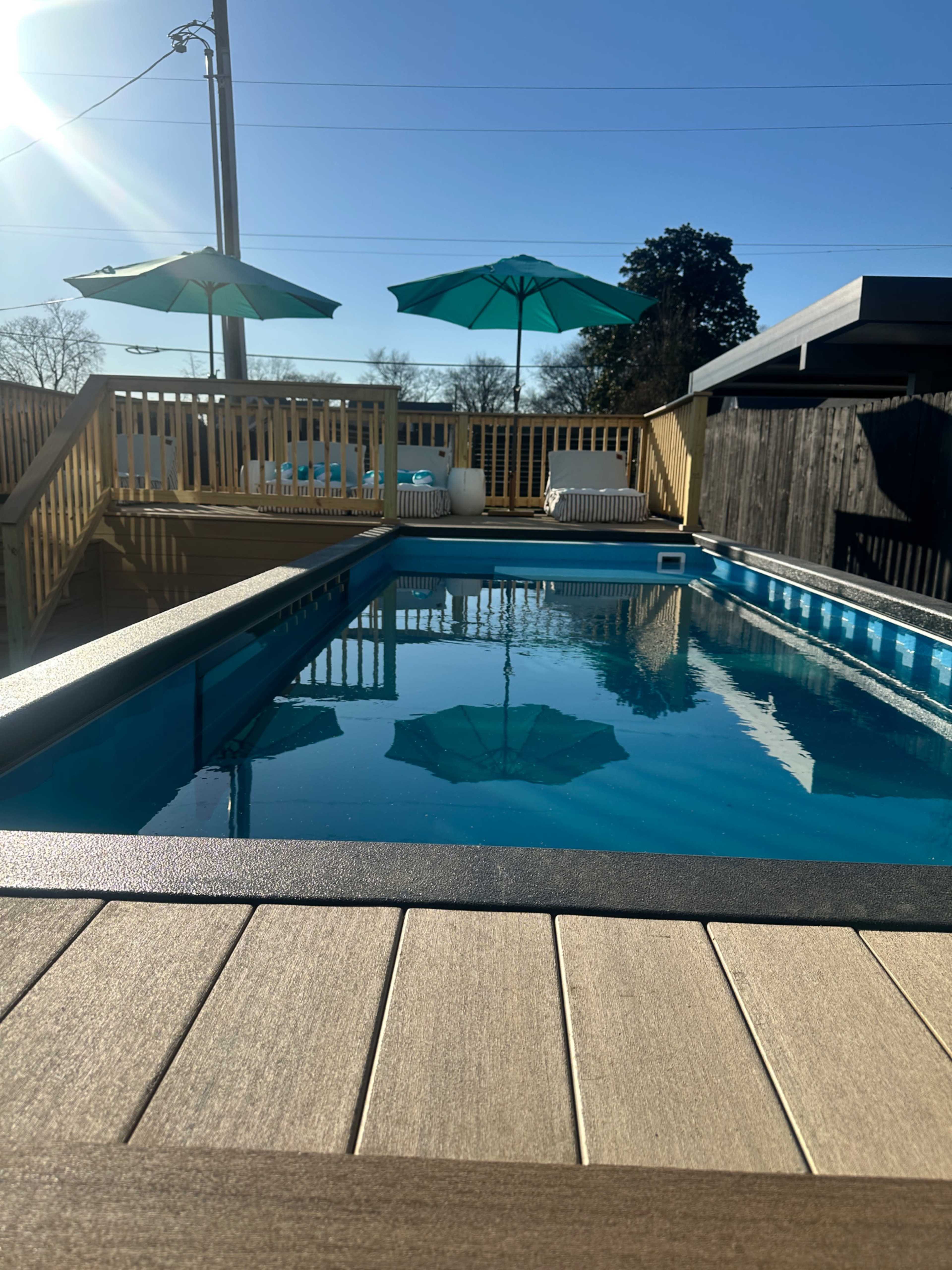 The image shows a swimming pool surrounded by wooden decking, with sun umbrellas in the background, beneath a clear blue sky.