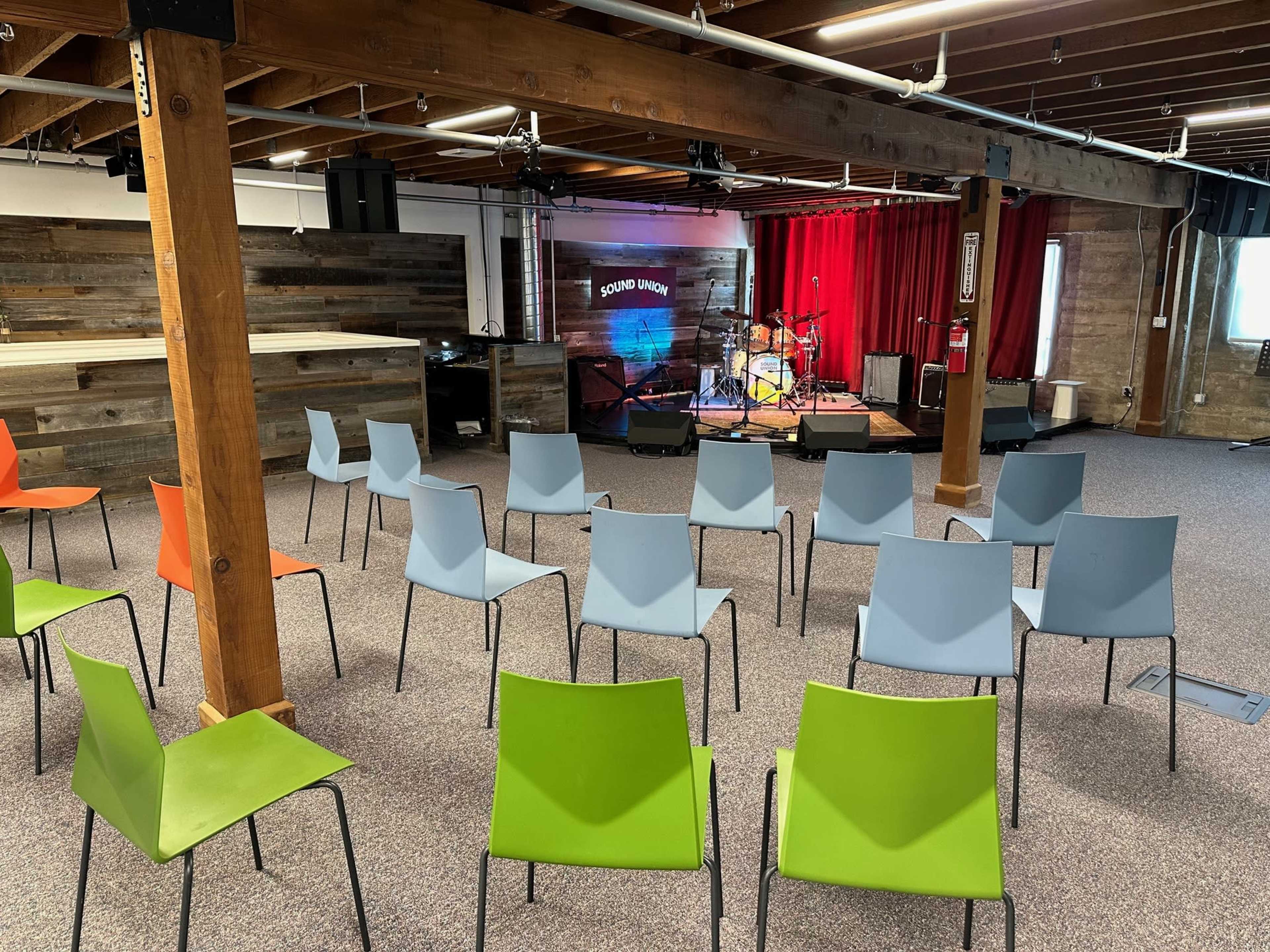 The image shows a small performance venue with empty colored chairs arranged in rows facing a stage, which is set up with musical instruments and backed by a red curtain.
