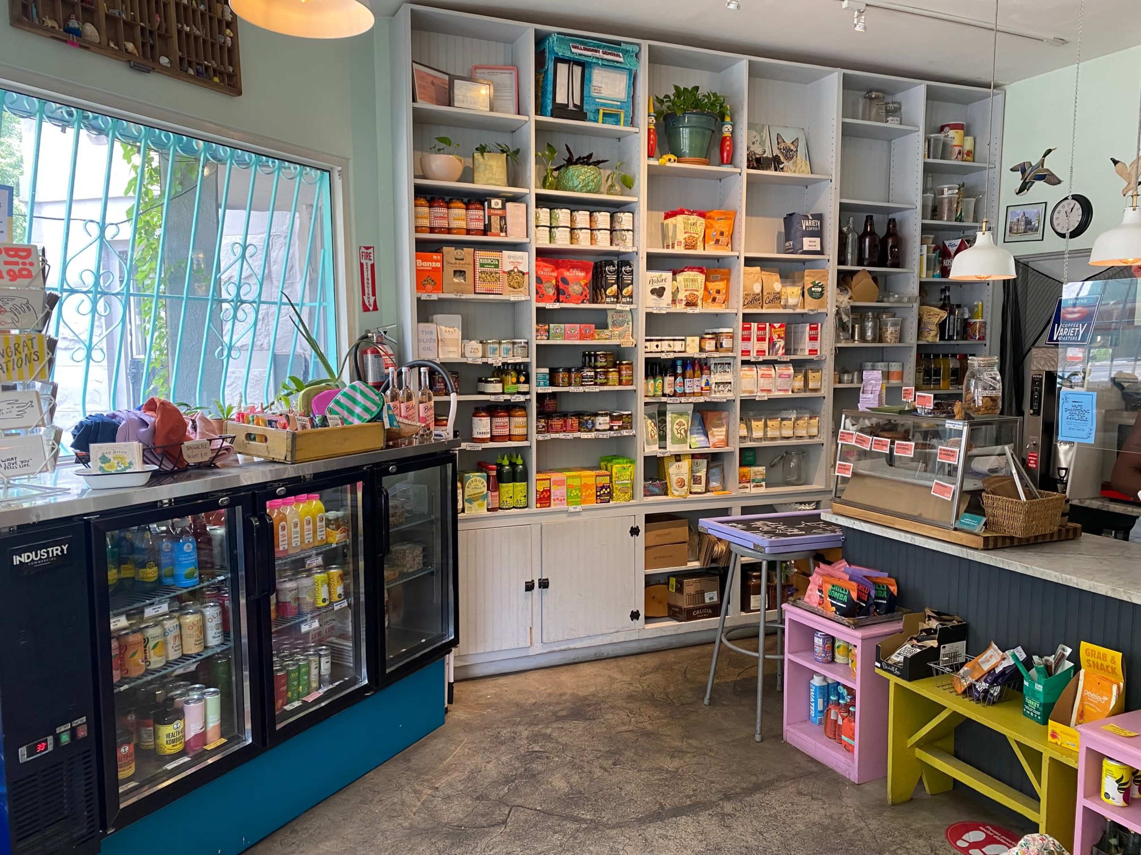 A small store interior featuring shelves stocked with various canned goods and products, a glass display refrigerator, and a counter with a cash register.