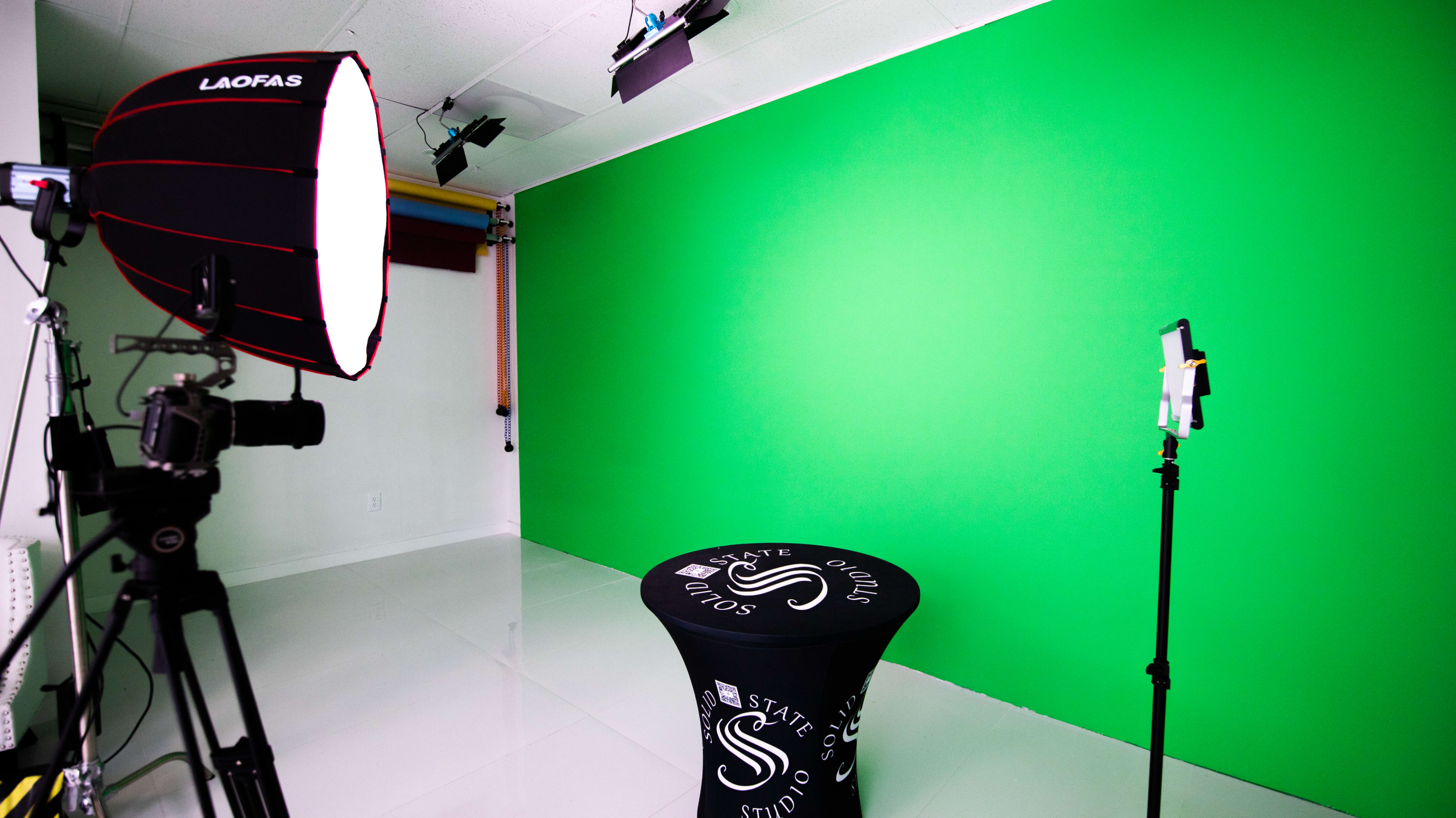 A studio setup featuring a green backdrop, a lighting umbrella, a camera on a tripod, and a table with a branded tablecloth.
