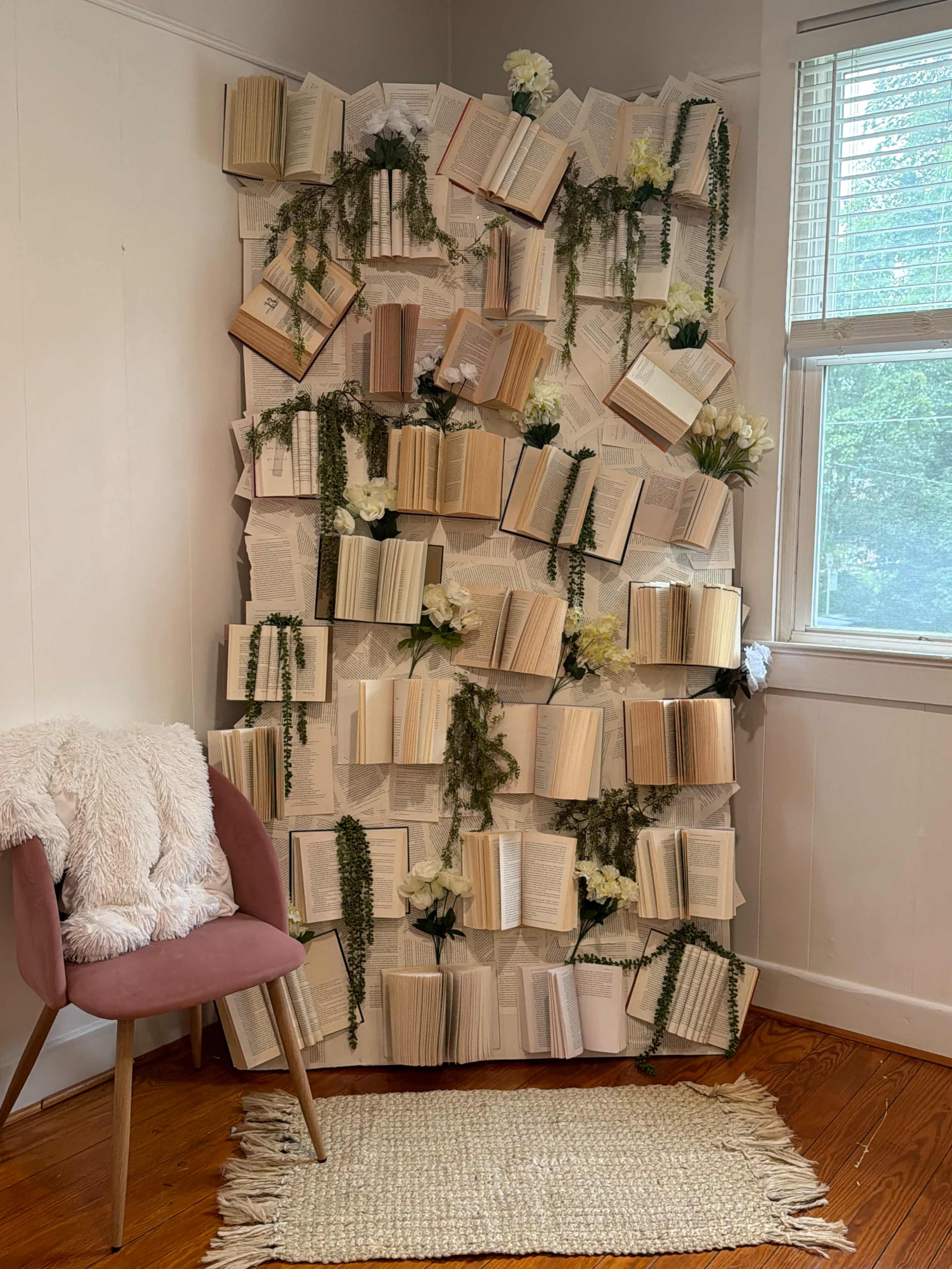 The image shows a decorative wall display made of open books arranged with greenery and flowers, alongside a pink chair and a woven rug on the wooden floor.