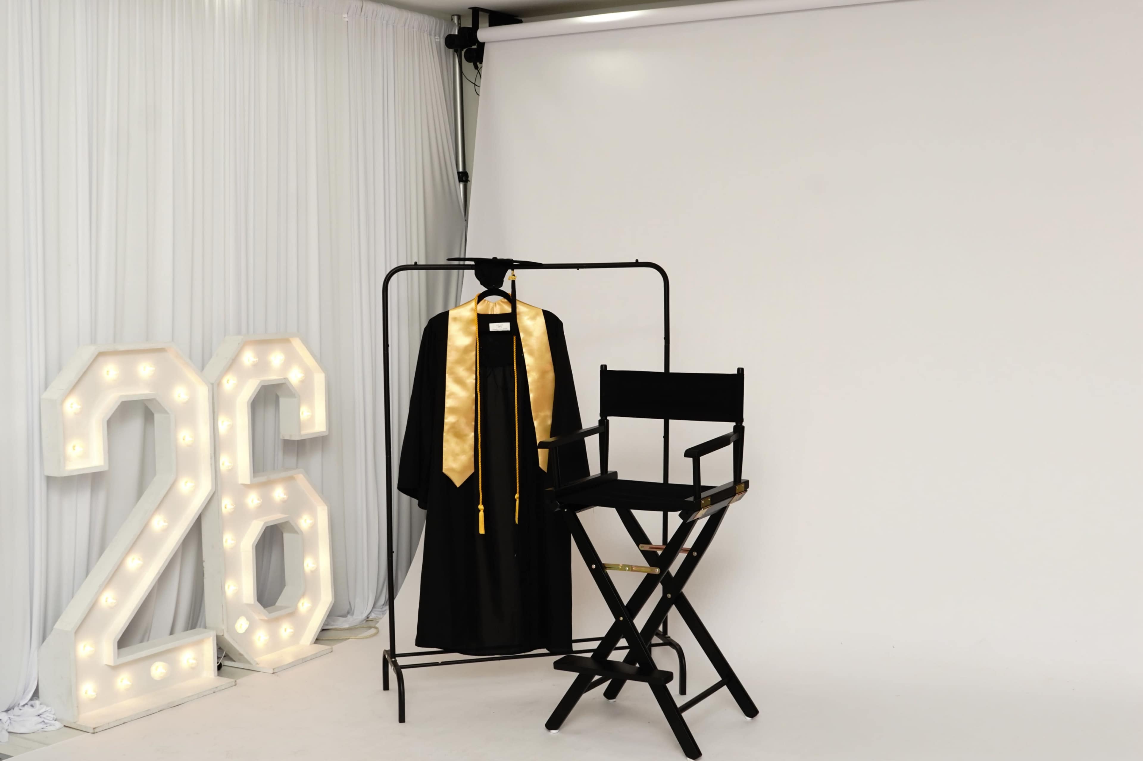 The image features a black graduation gown with gold accents hanging on a rack beside a black director's chair and illuminated numbers reading "26" against a plain white backdrop.