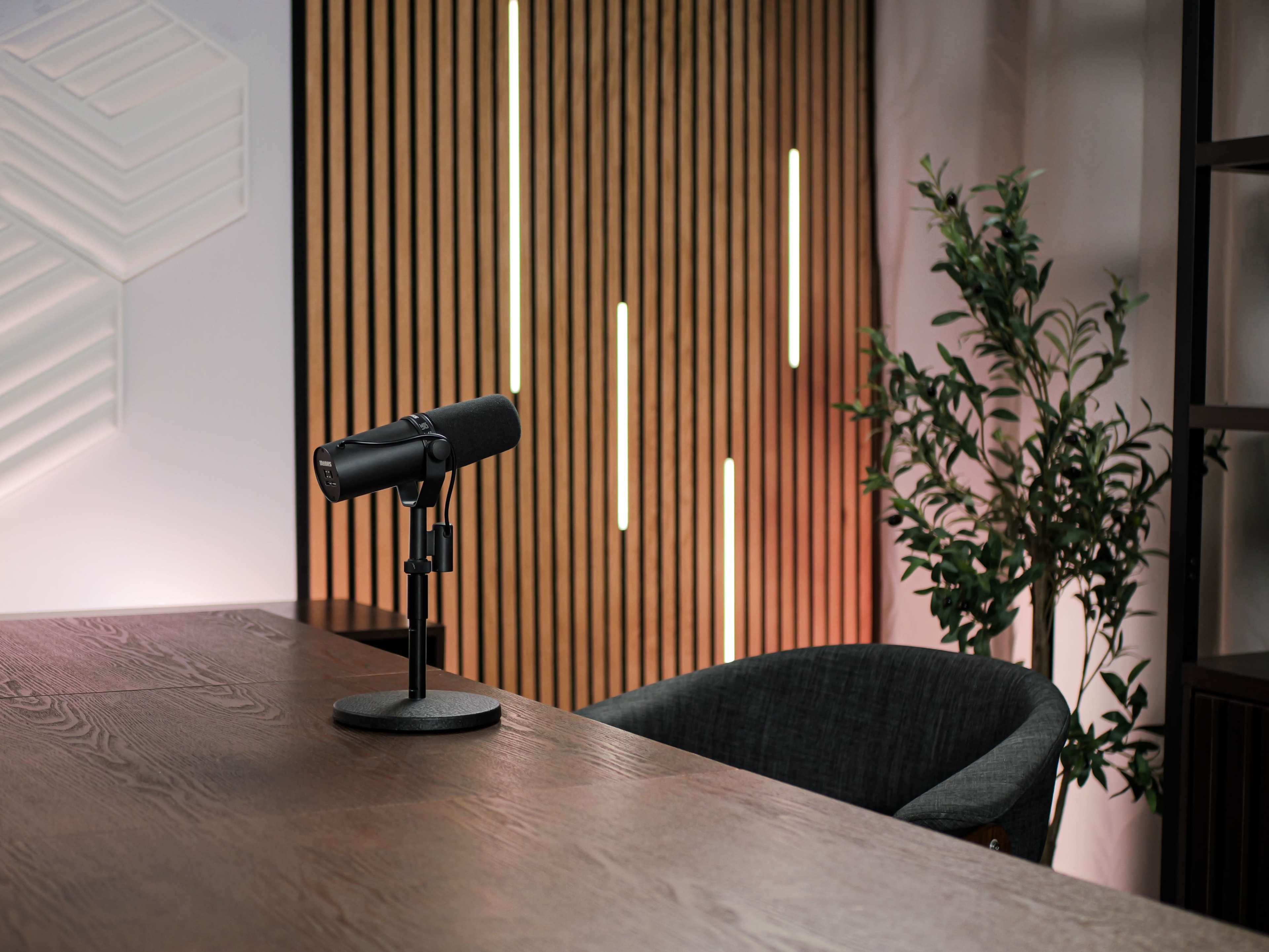 A microphone is positioned on a wooden table beside a modern chair and a decorative plant, against a backdrop of vertical wooden slats and ambient lighting.
