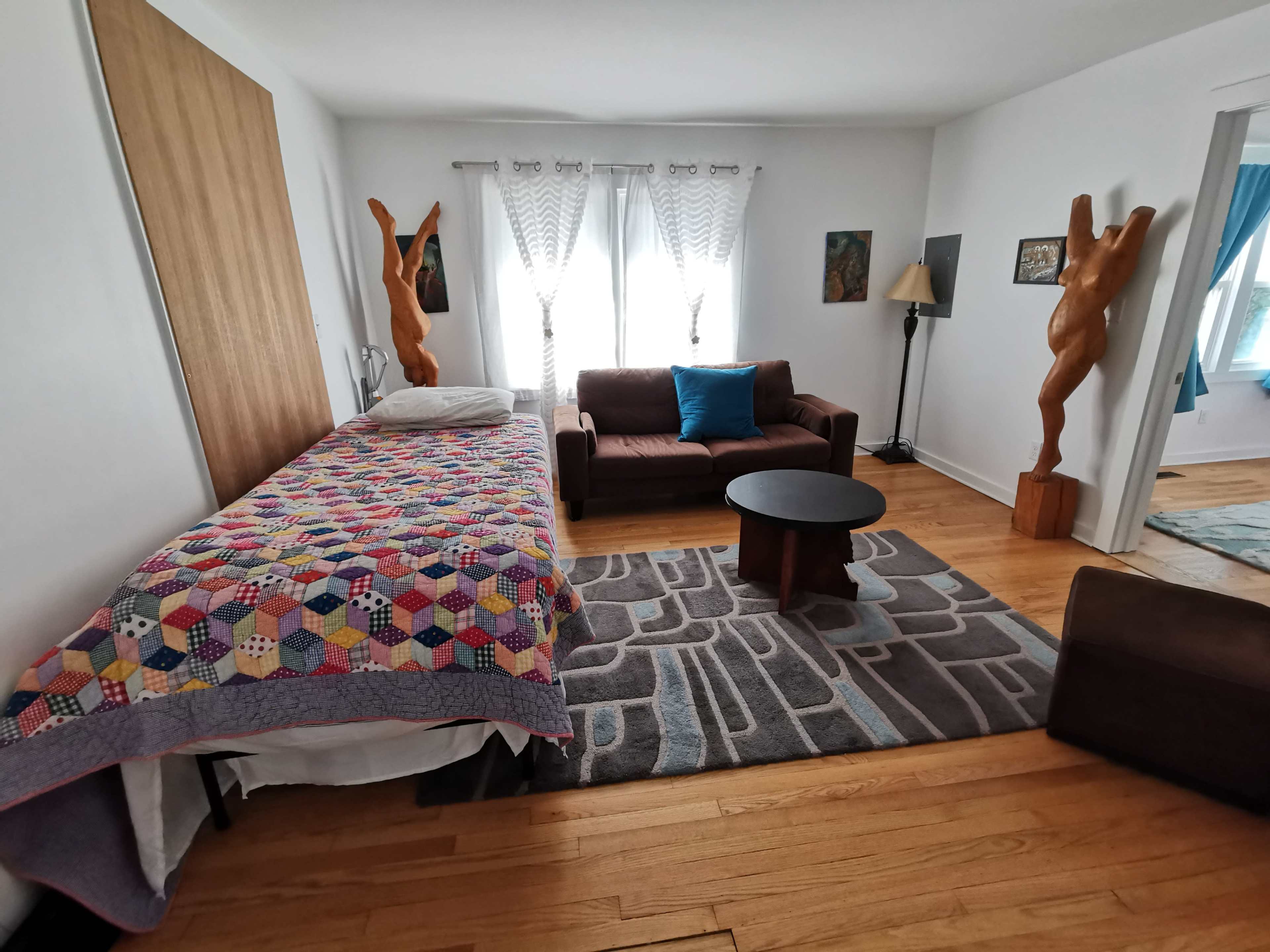 The room features a bed covered with a colorful quilt, a brown couch with a blue pillow, a round table, and two wooden sculptures near the windows.