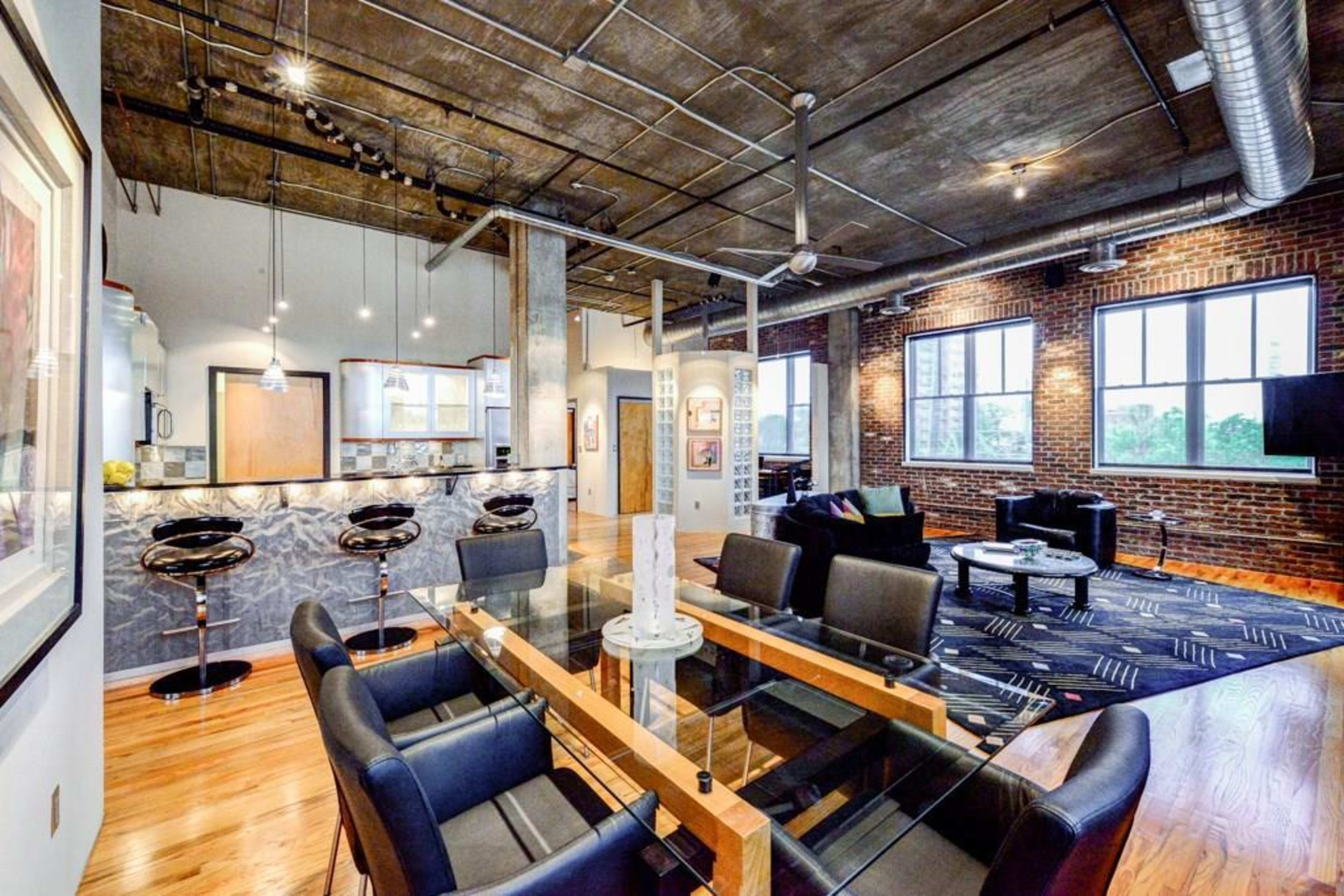 A modern loft-style living space featuring an open layout with exposed brick walls, hardwood floors, and a glass dining table surrounded by black chairs.