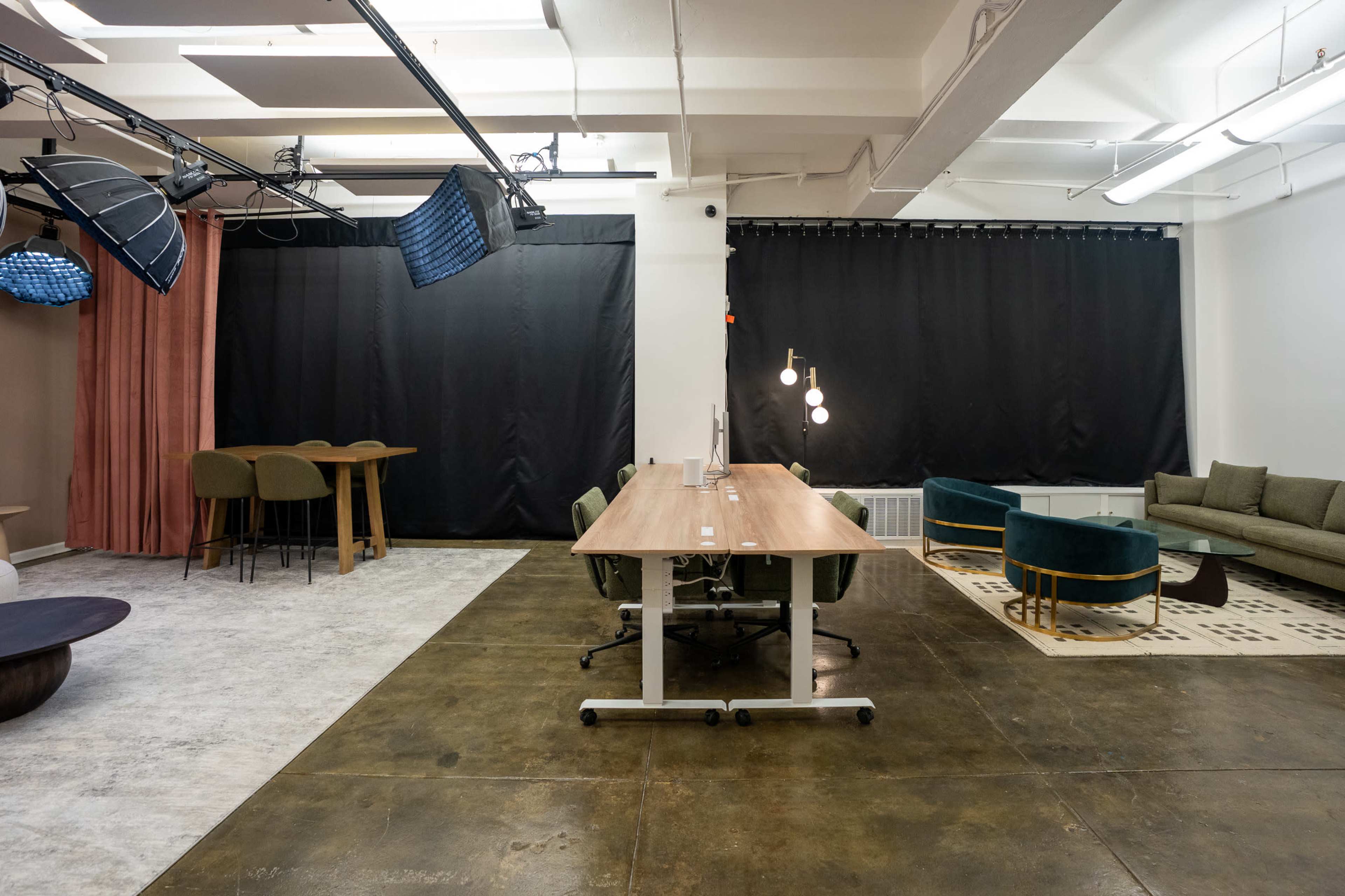 The image shows a modern office space featuring a long wooden table with chairs, a seating area with green chairs and a sofa, and various lighting fixtures.