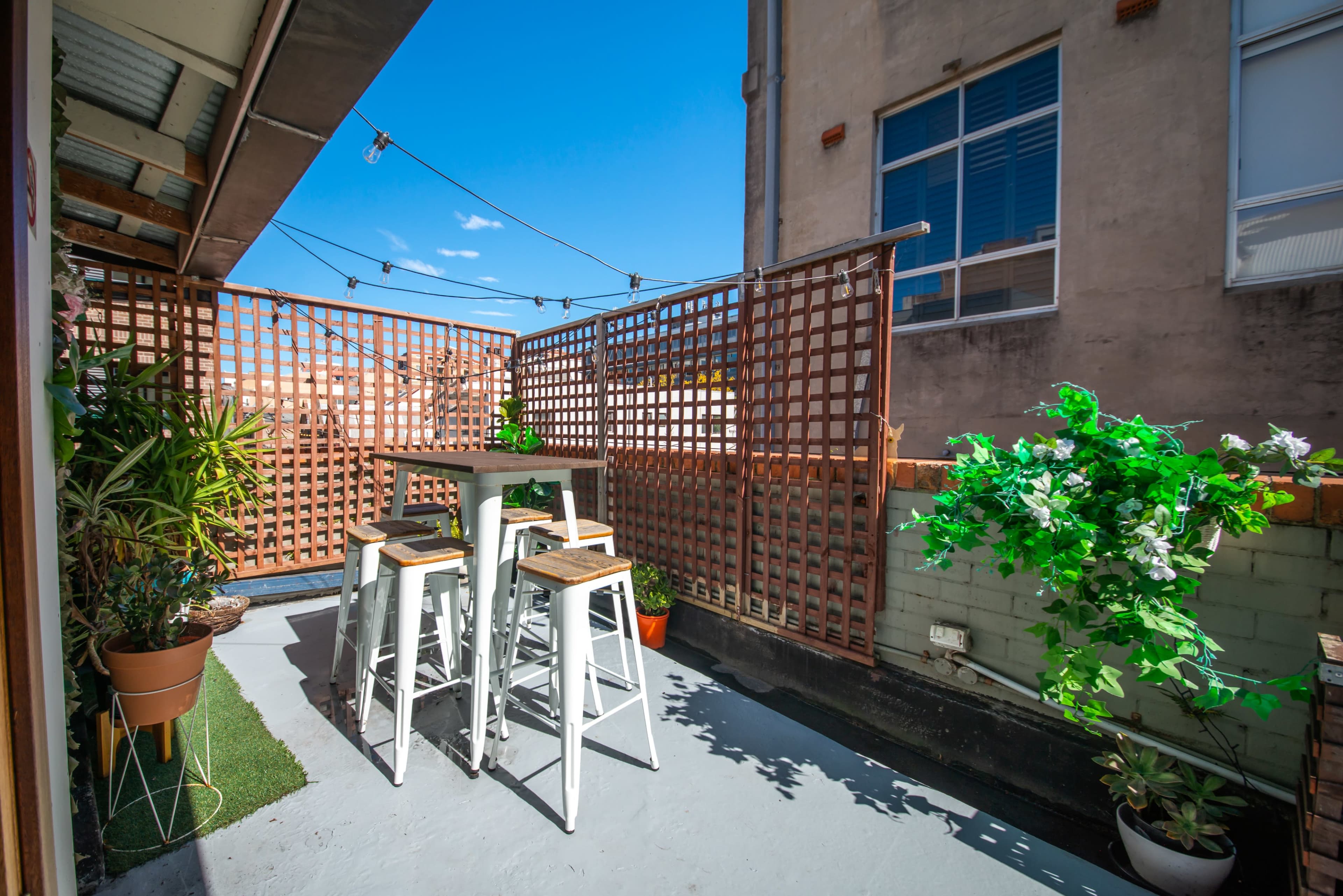 A small outdoor space features a table with four high stools, surrounded by lattice screens and potted plants under a clear blue sky.