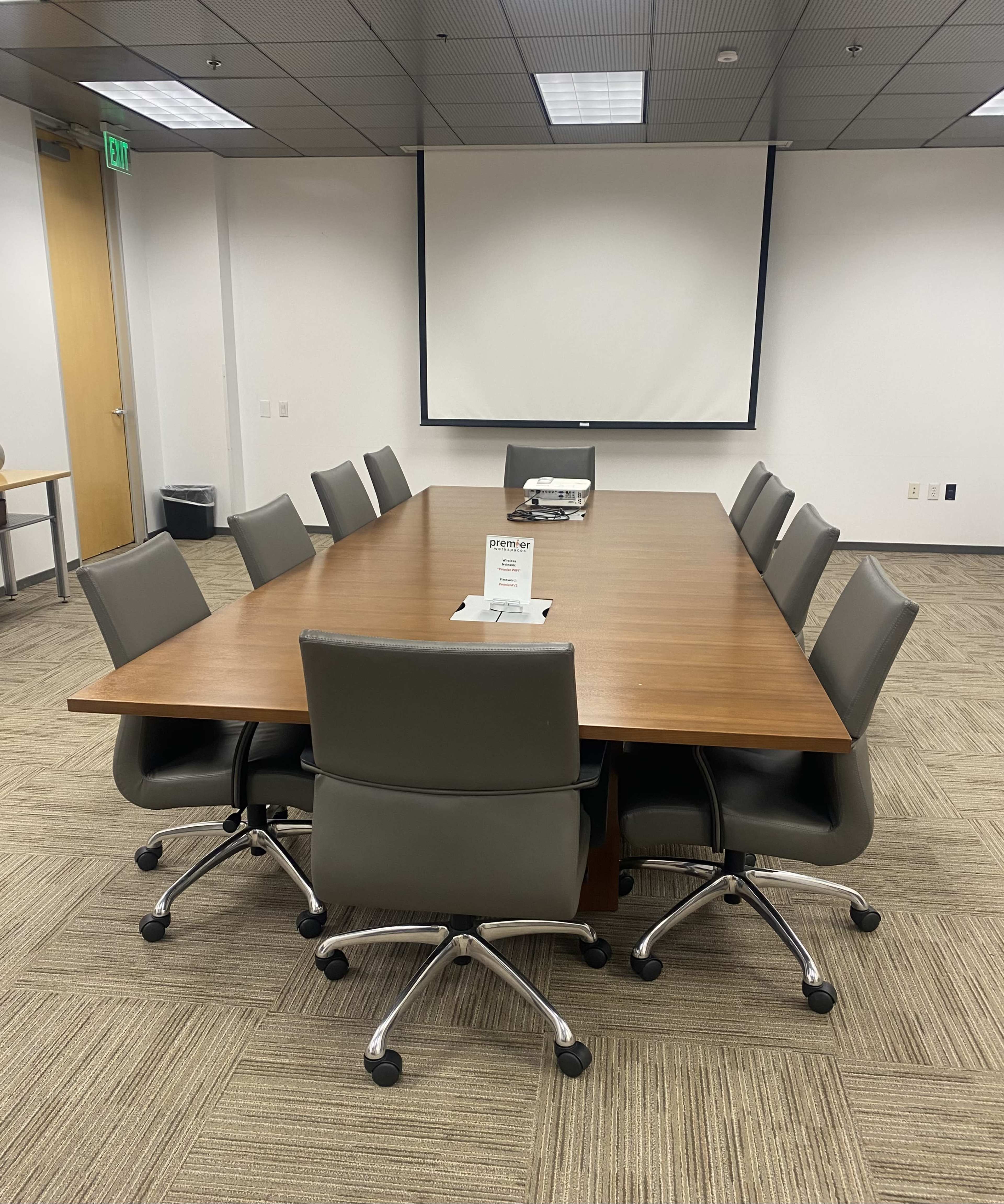 A large conference room is set up with a long wooden table surrounded by eight ergonomic chairs, and a projector screen is visible at one end.