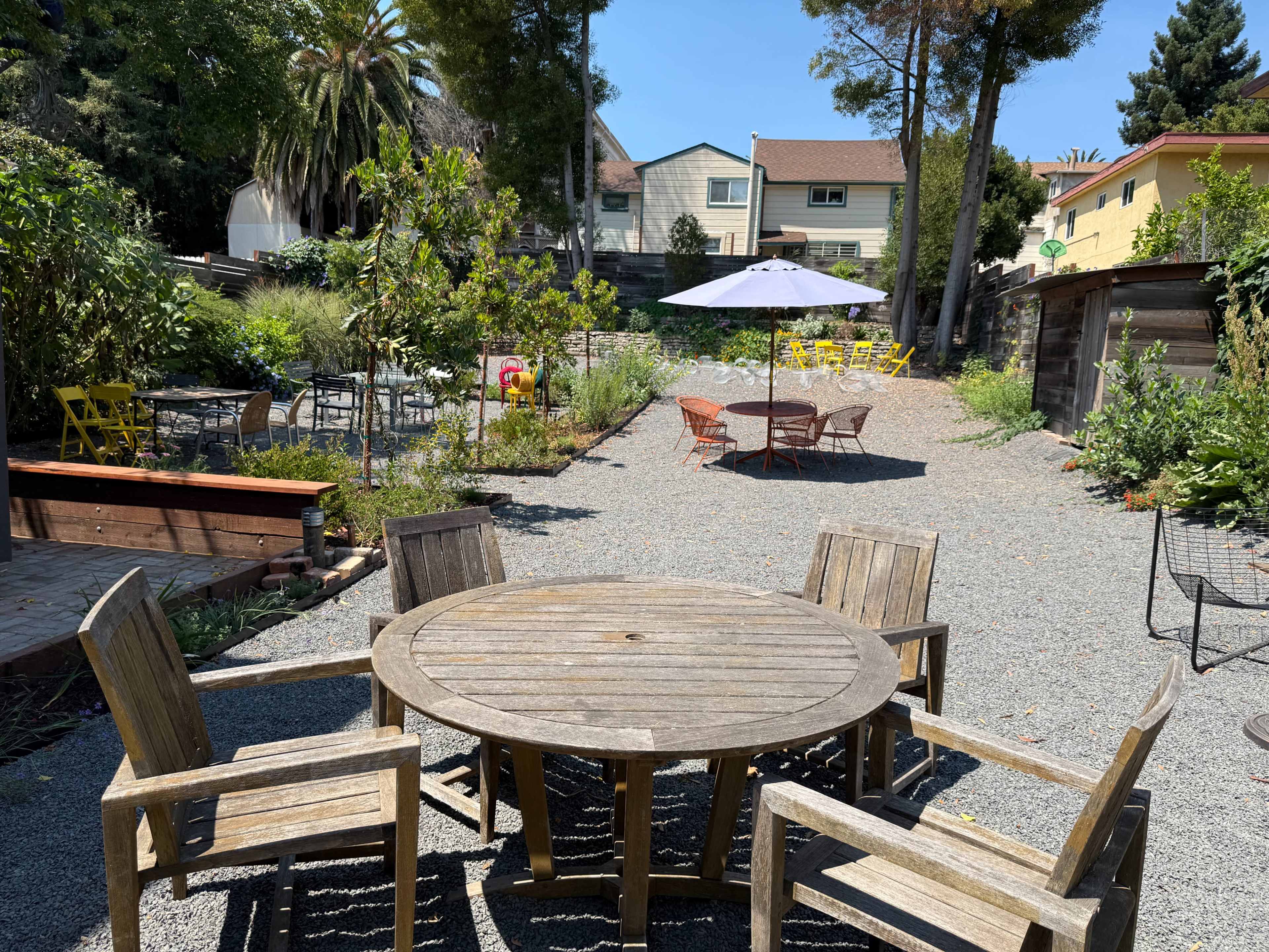 The image shows a garden area with a wooden table and chairs surrounded by gravel, colorful chairs, and greenery under a clear blue sky.