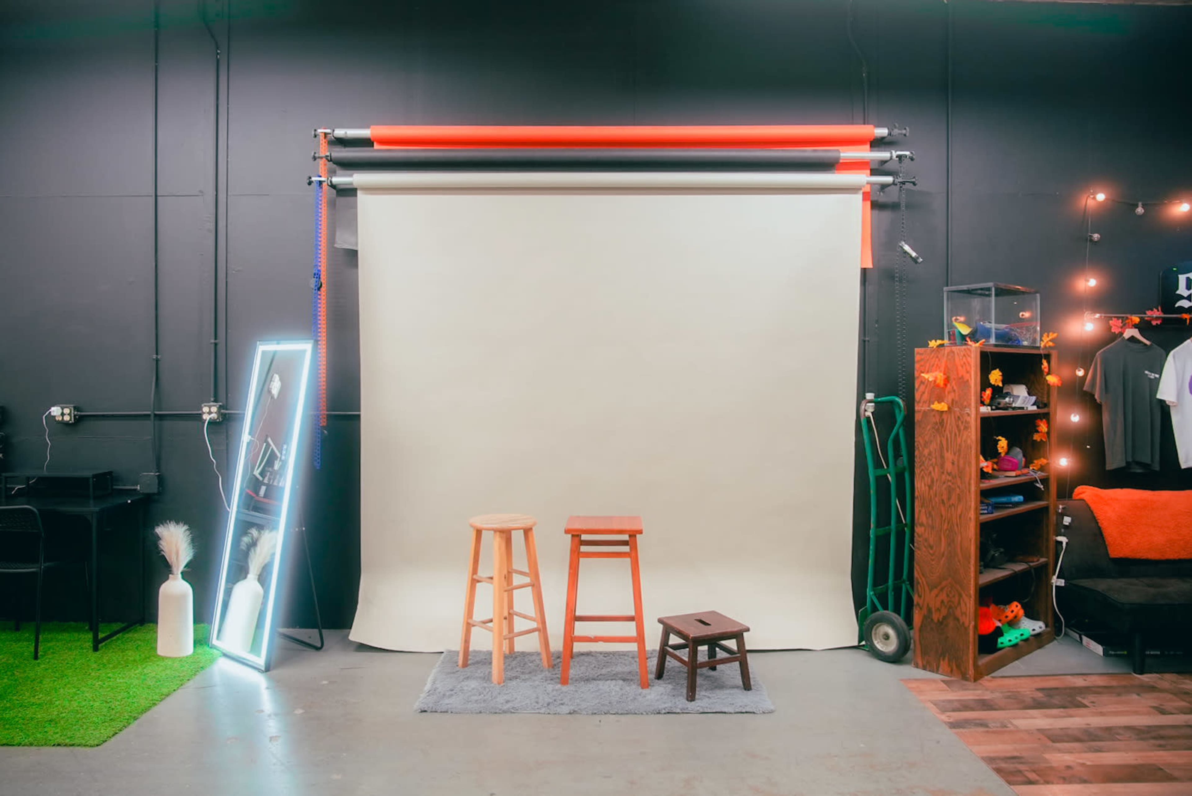 A studio setup with a plain backdrop, three stools of varying heights in front, and a shelving unit displaying various items to the side.