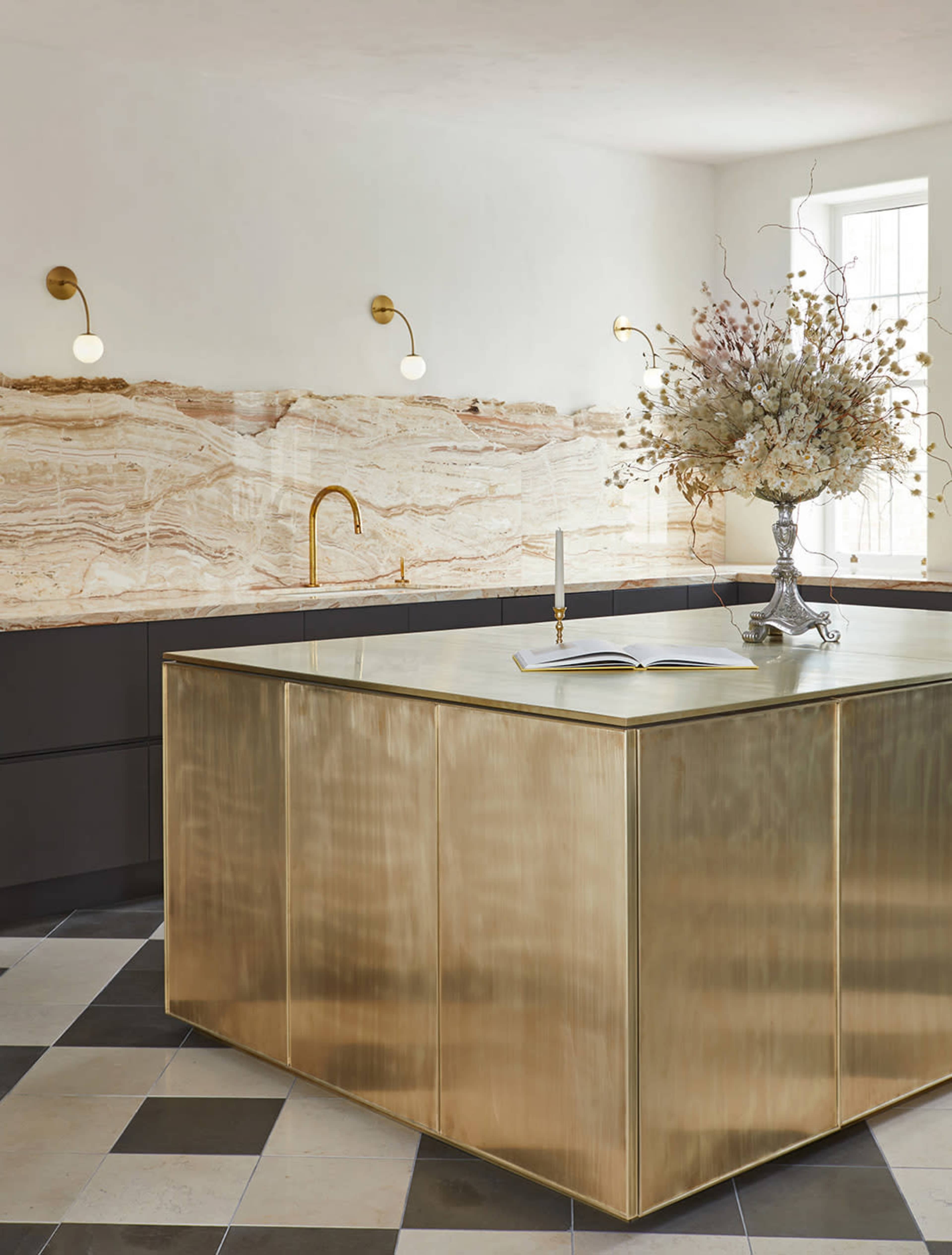 The image features a kitchen with a large brass island, dark cabinetry, and a stone wall backsplash, accented by a floral arrangement in a decorative vase.