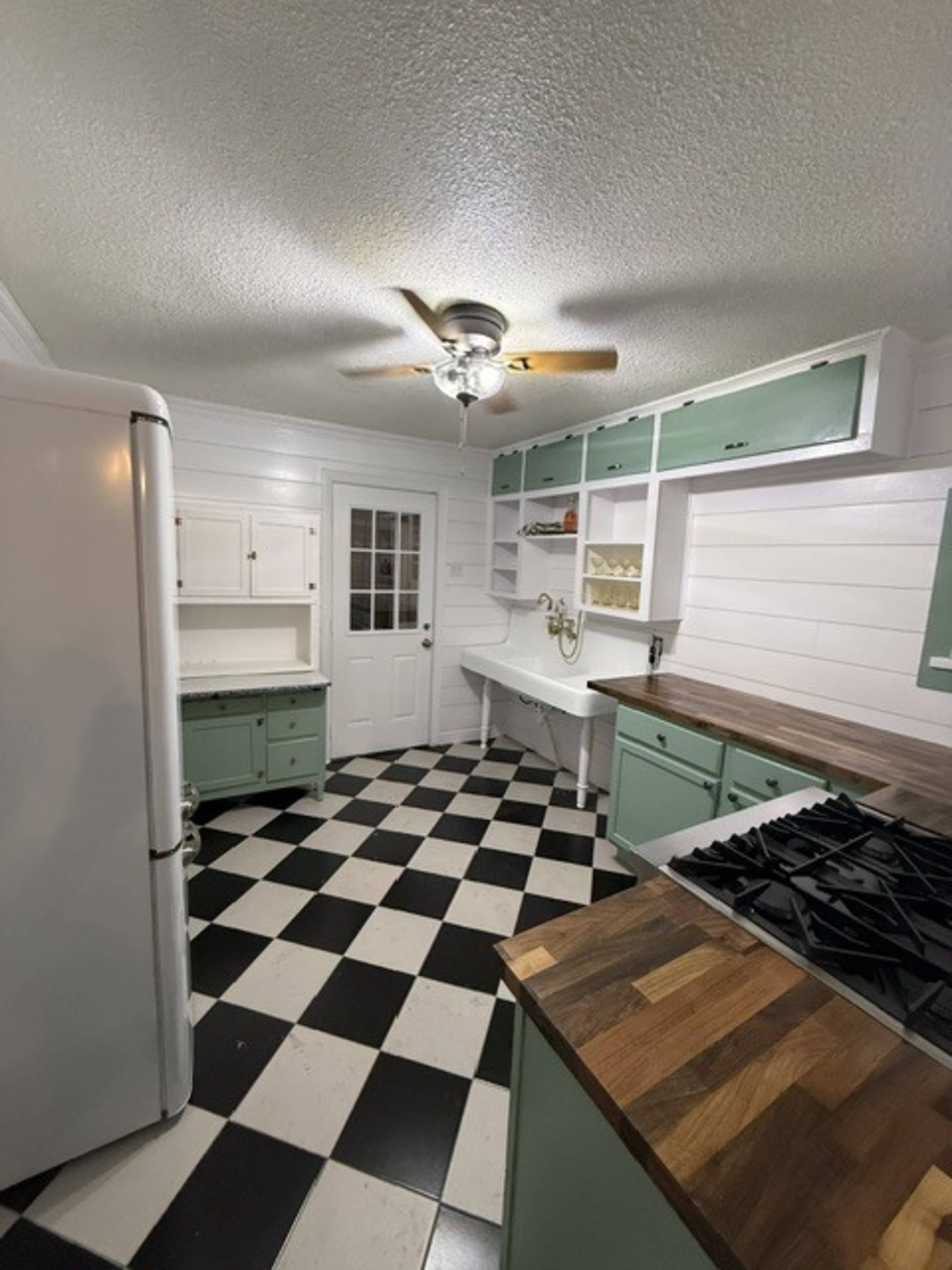The image shows a kitchen with a checkered black and white floor, mint green cabinetry, a wooden countertop, and a ceiling fan.