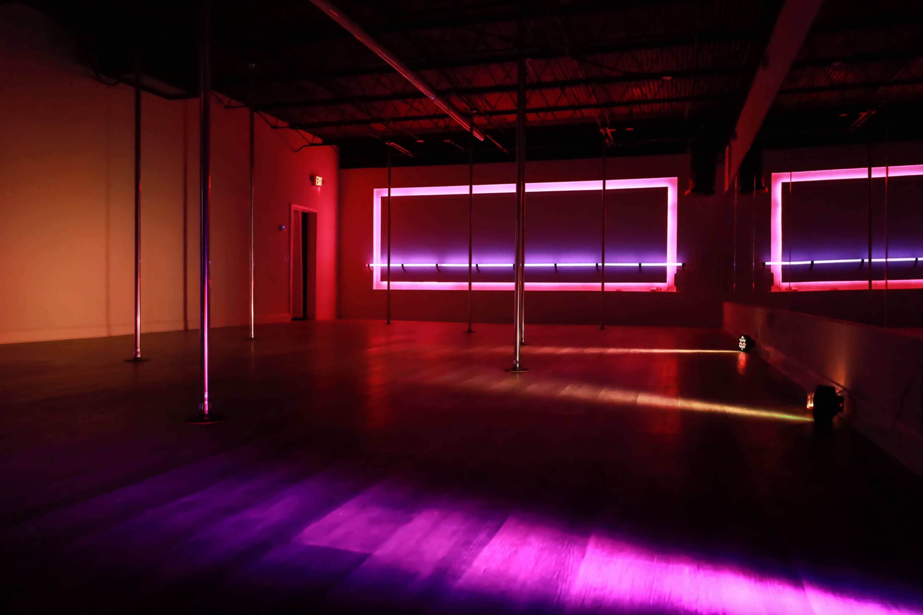 The image shows an empty dance studio with mirrored walls, featuring several poles and illuminated with purple and pink lighting.