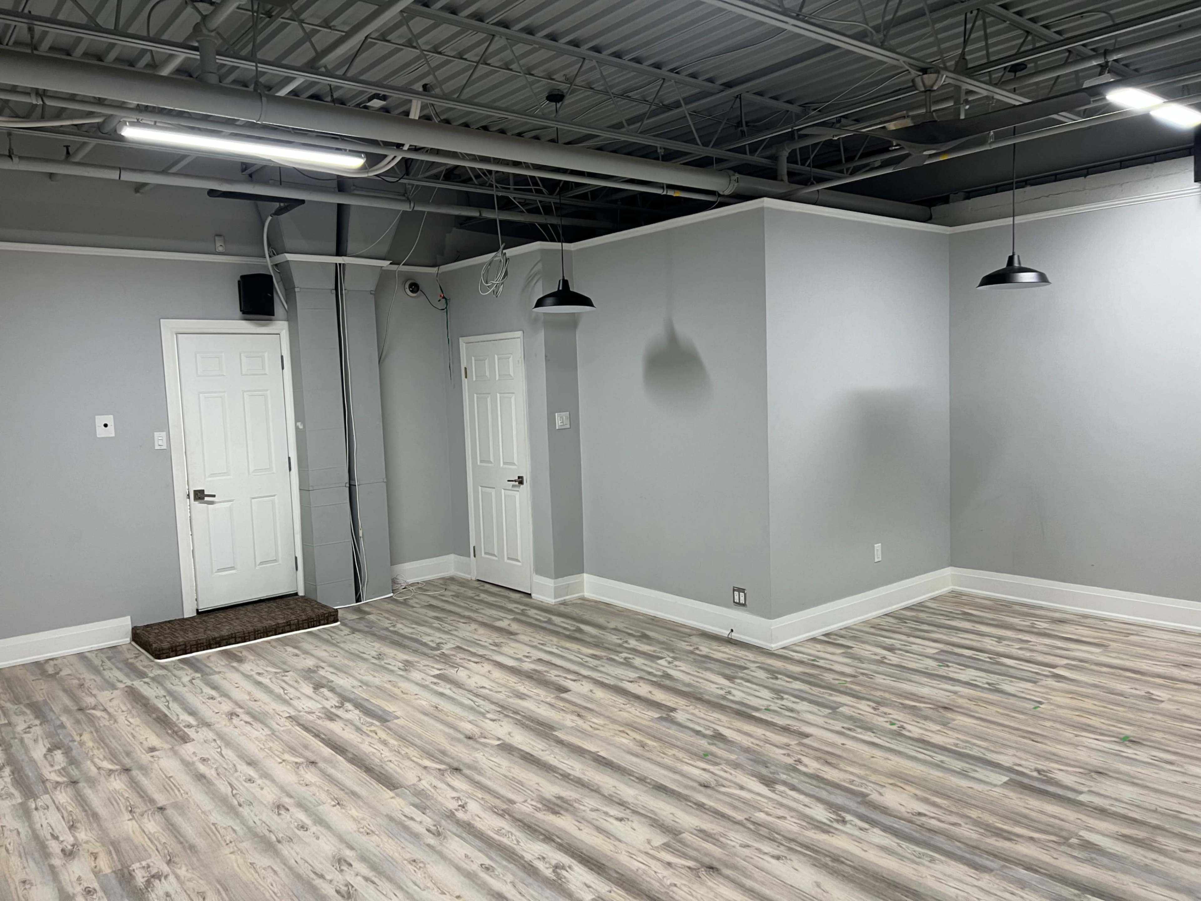 A partially empty, light gray room with two doors, hanging lights, and wooden flooring.