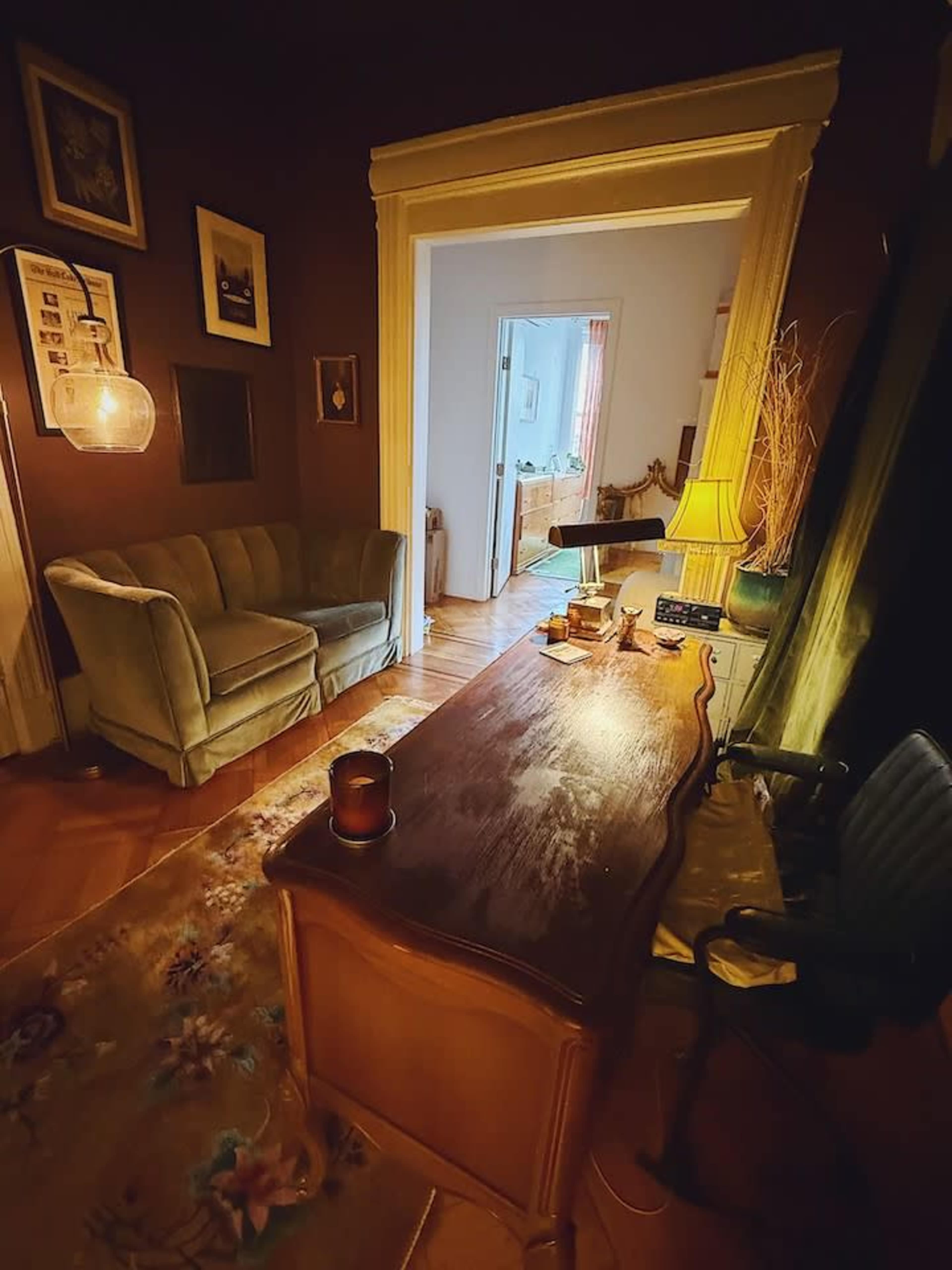 The image shows a cozy room featuring a wooden desk, a green couch, and framed pictures on the walls, with a doorway leading to another space in the background.