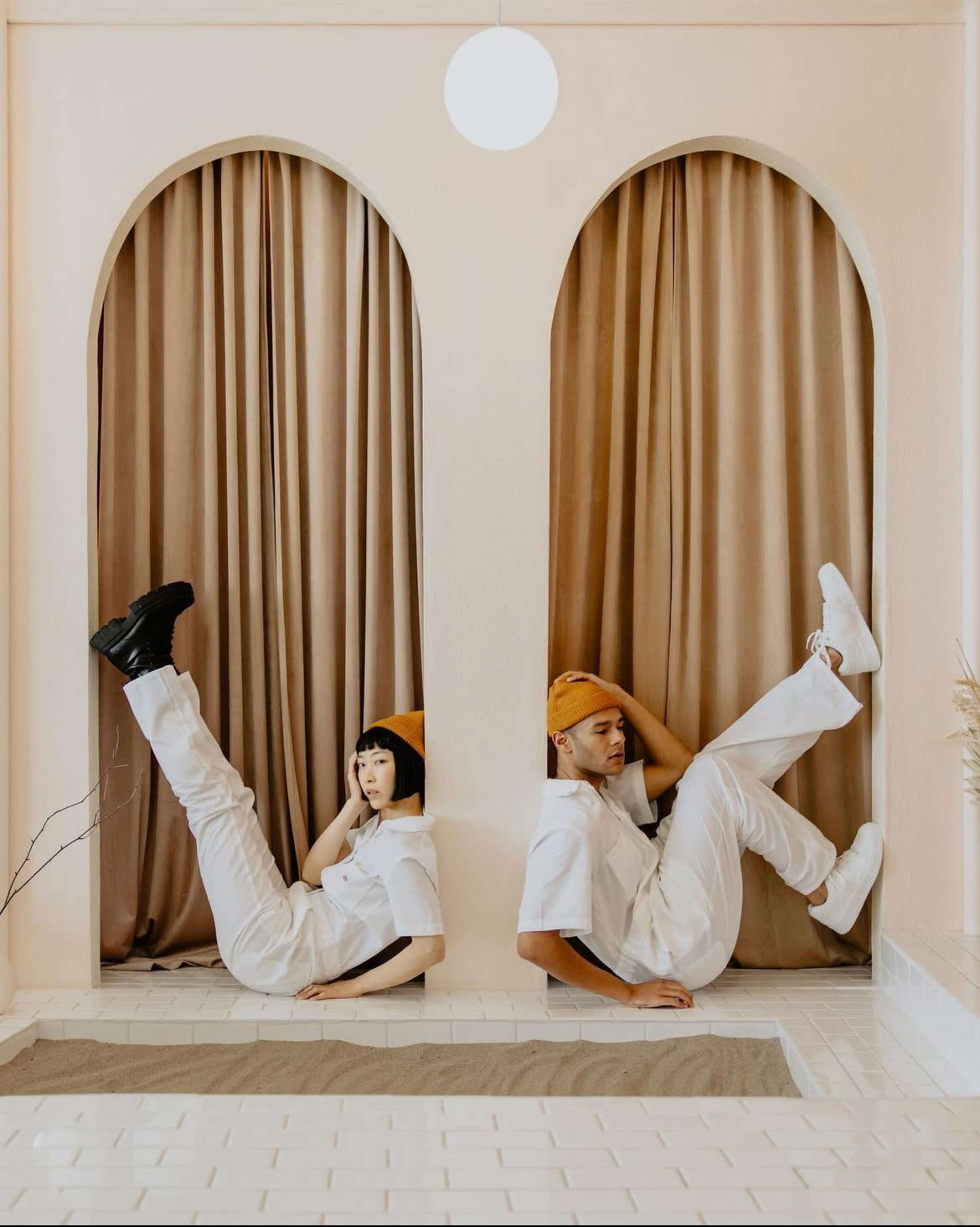 A man and a woman in white outfits pose next to each other, leaning against a wall framed by arched curtains, with their legs raised in the air.