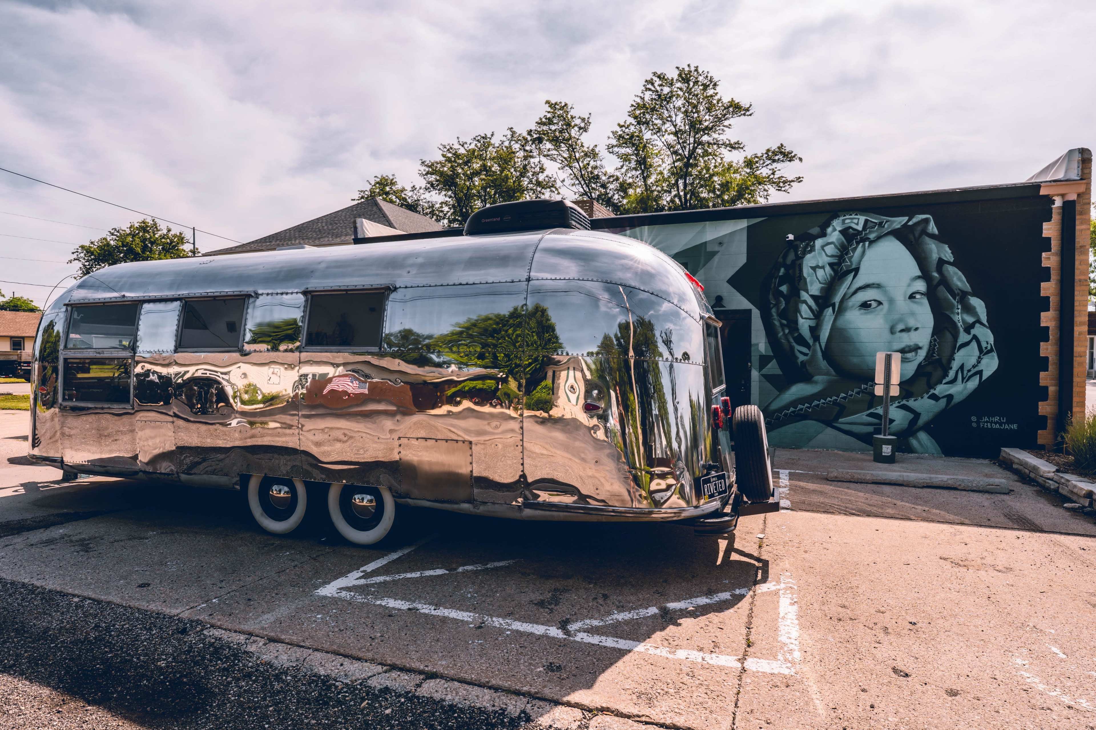 A shiny, silver Airstream trailer is parked next to a mural depicting a woman with a headscarf.