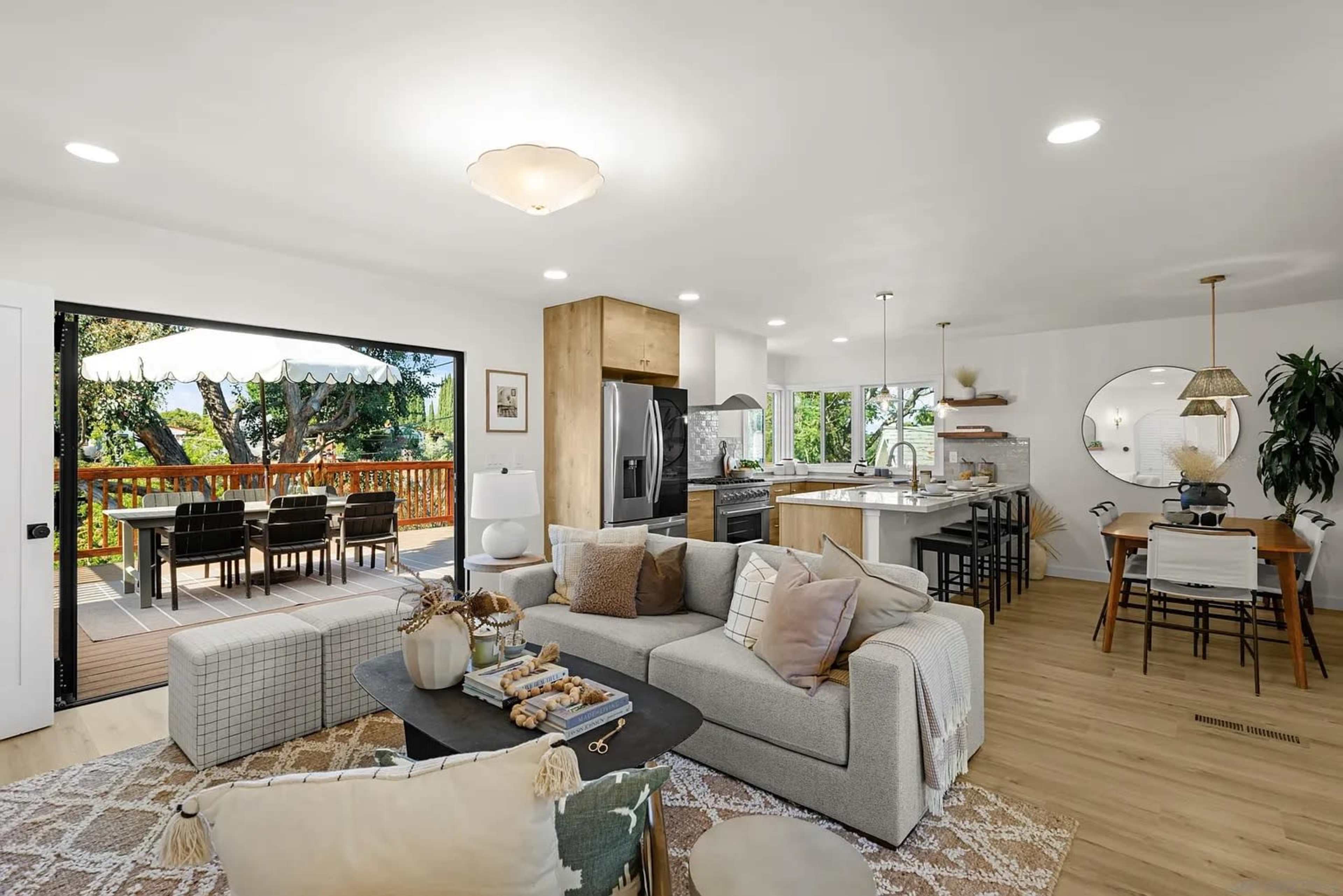 The image shows a modern open-concept living space with a living area, dining area, and kitchen, featuring large windows that open to a deck.