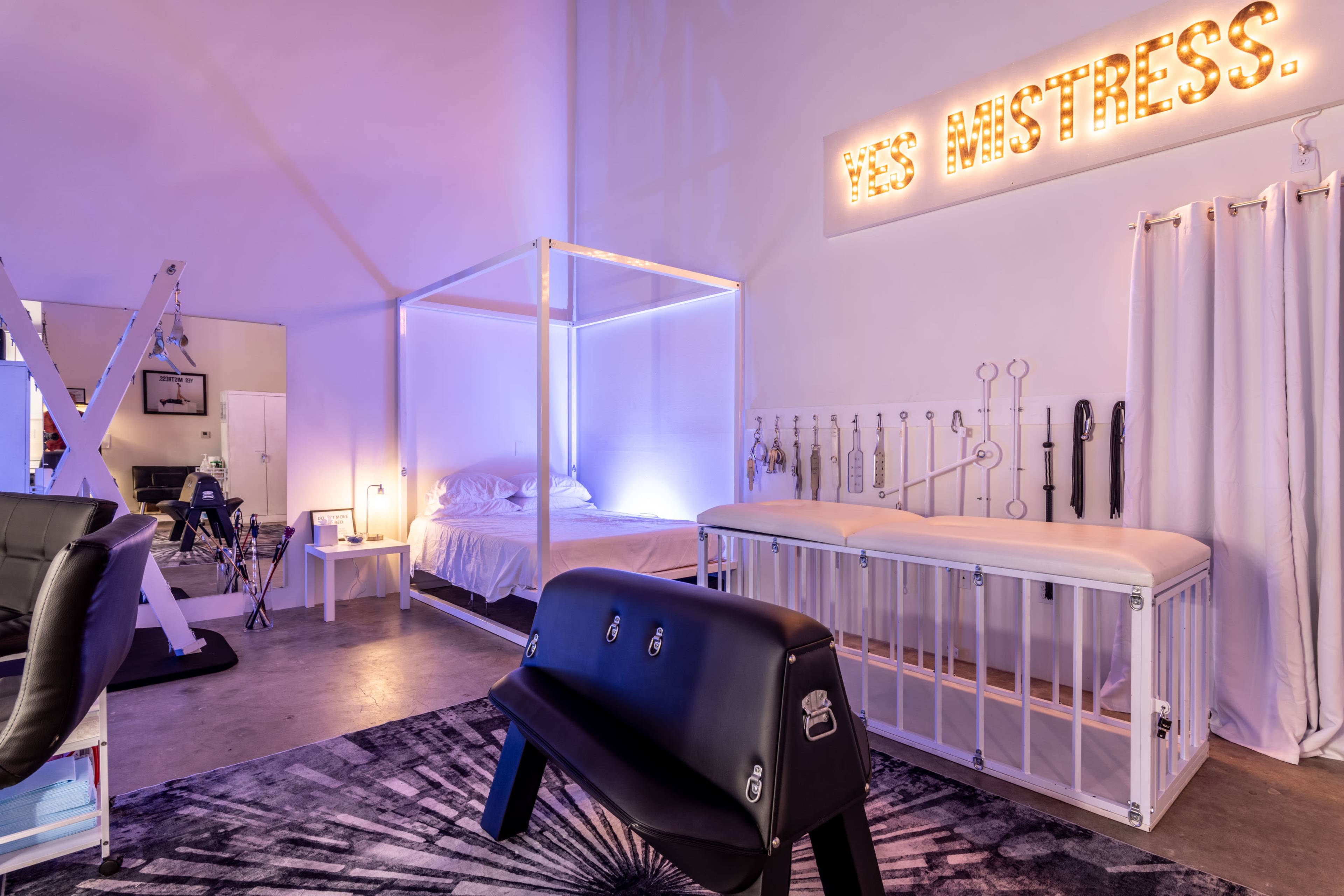 The image depicts a room designed for adult-themed activities, featuring a bed with a canopy, various tools mounted on the wall, and a bench in the center.