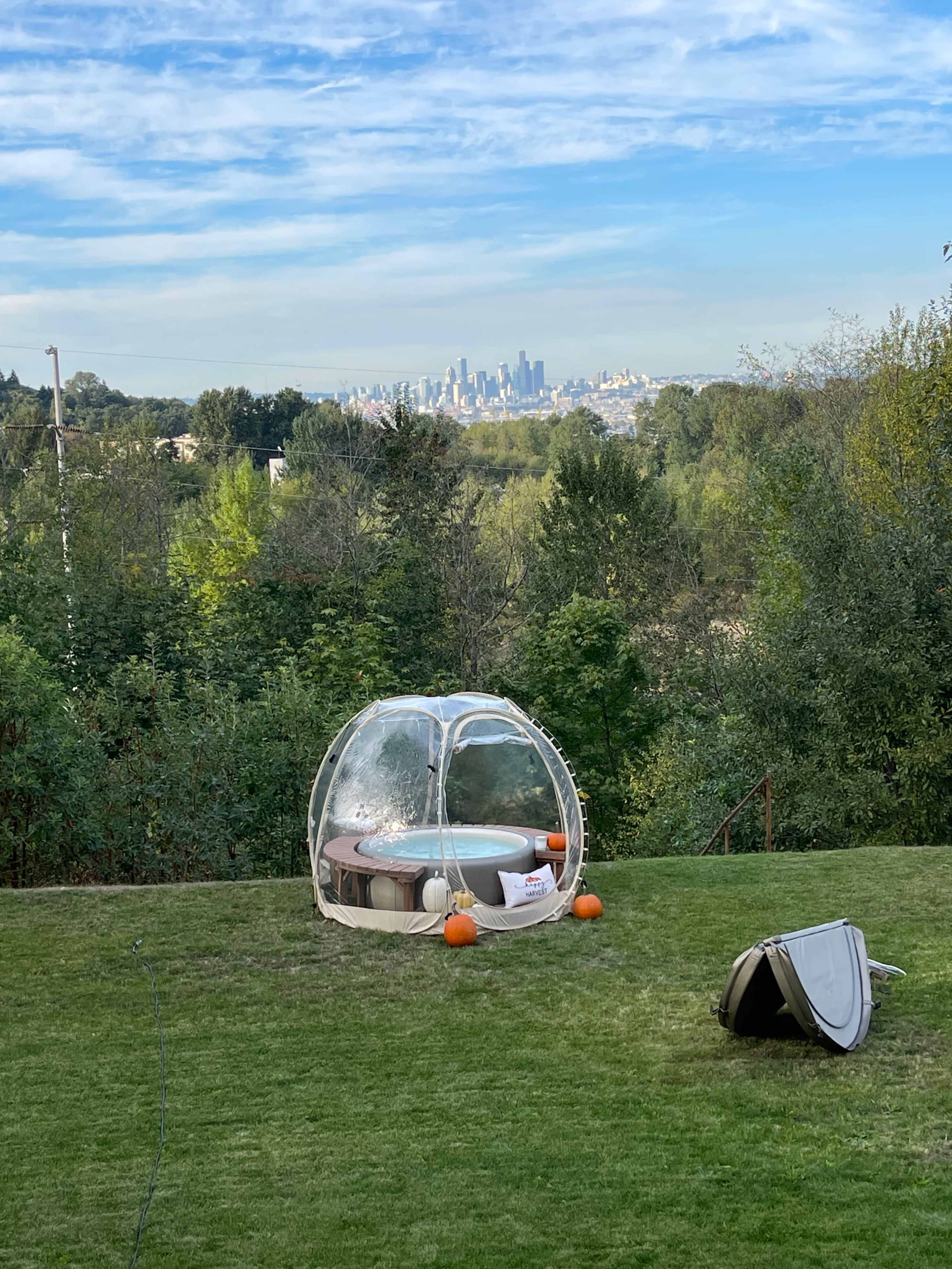 A clear bubble structure is set up on a grassy area with pumpkins nearby, overlooking a city skyline in the distance.