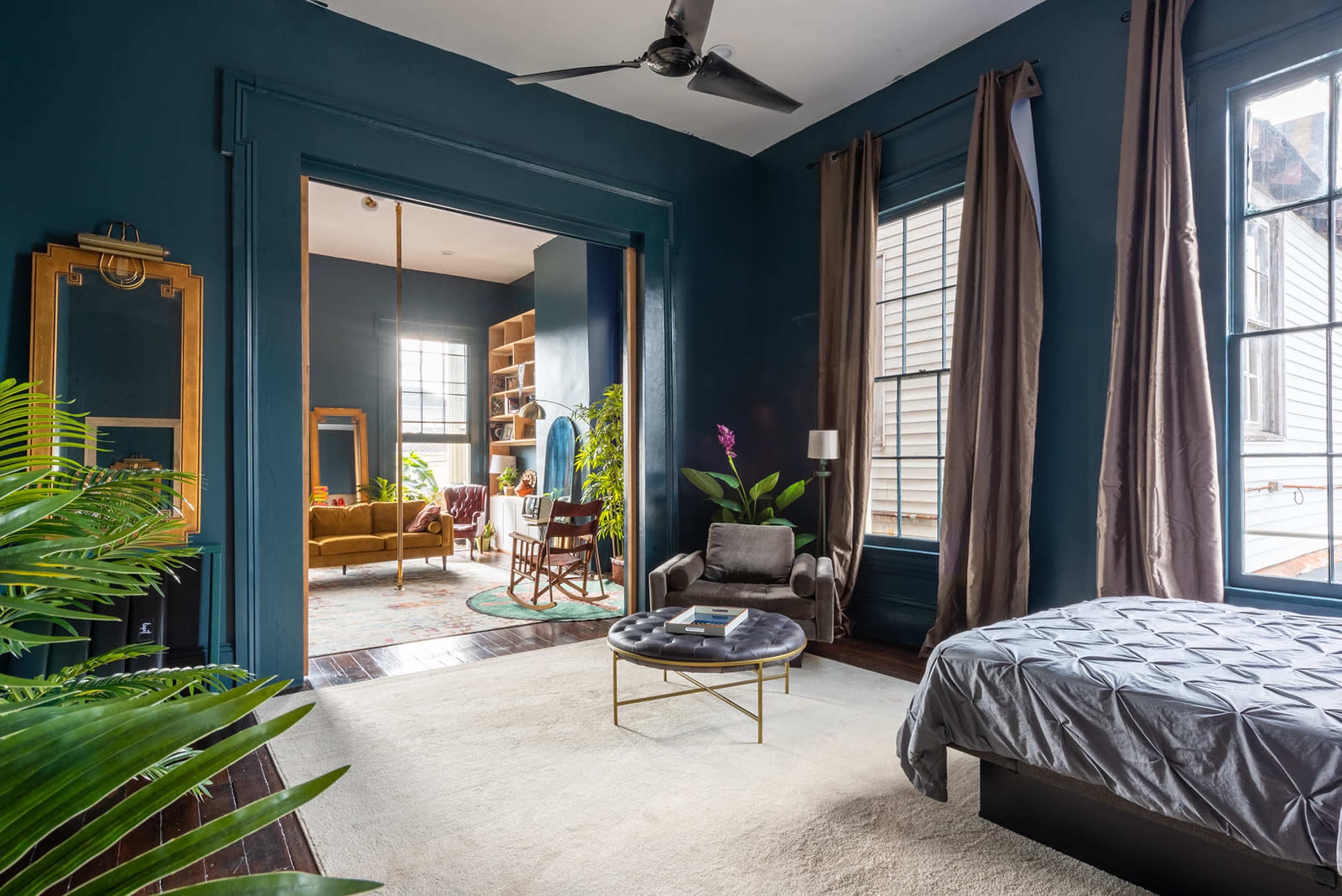 A master bedroom features a gray bedspread, a round coffee table, and large windows with plants in the foreground and an adjoining room visible through an open doorway.