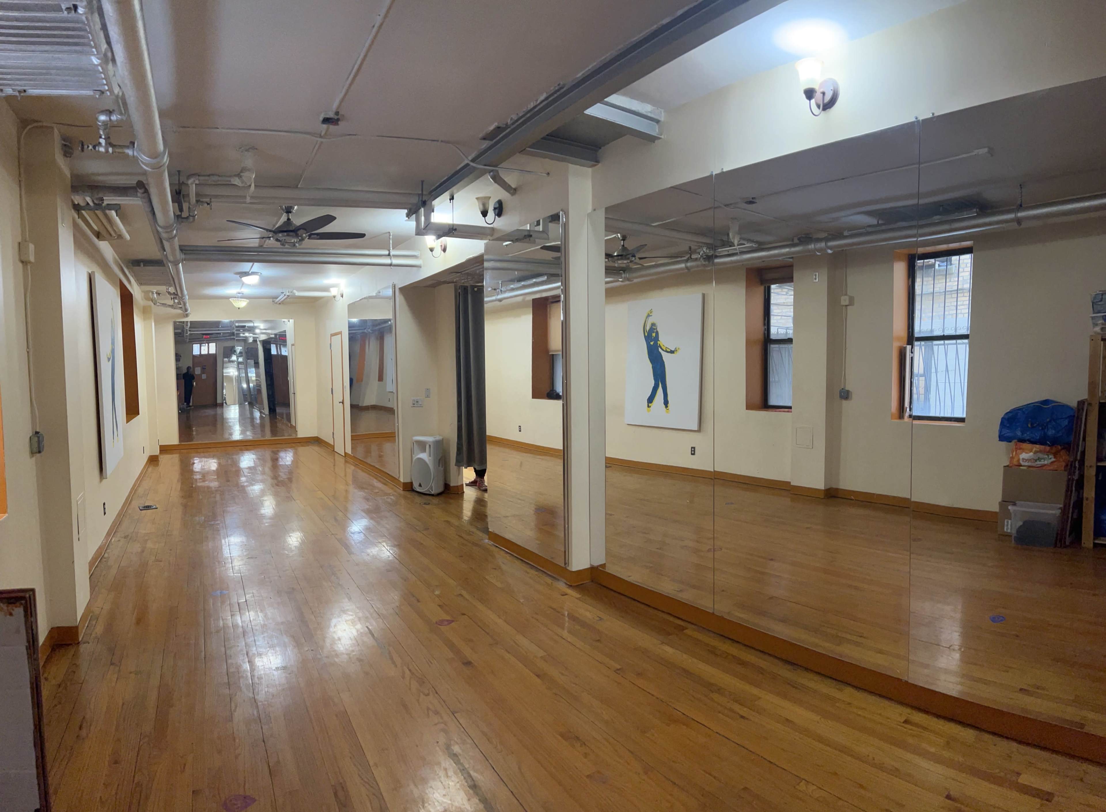 A spacious, well-lit room with polished wooden floors, featuring mirrors along one wall and a mural of a dancer.