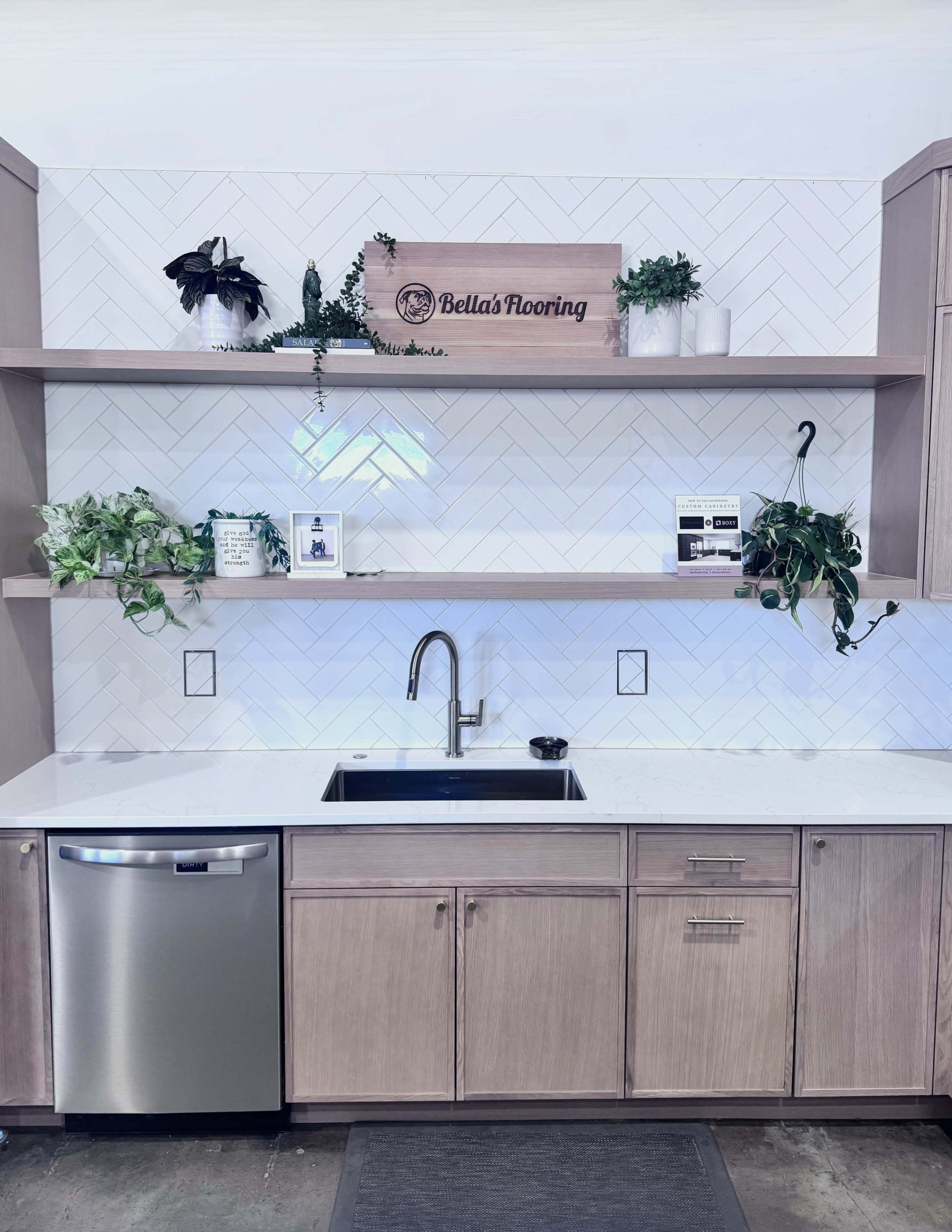 The image shows a modern kitchen with a stainless steel dishwasher, a deep sink, and open shelves adorned with plants and decorative items above white, herringbone-patterned tiles.