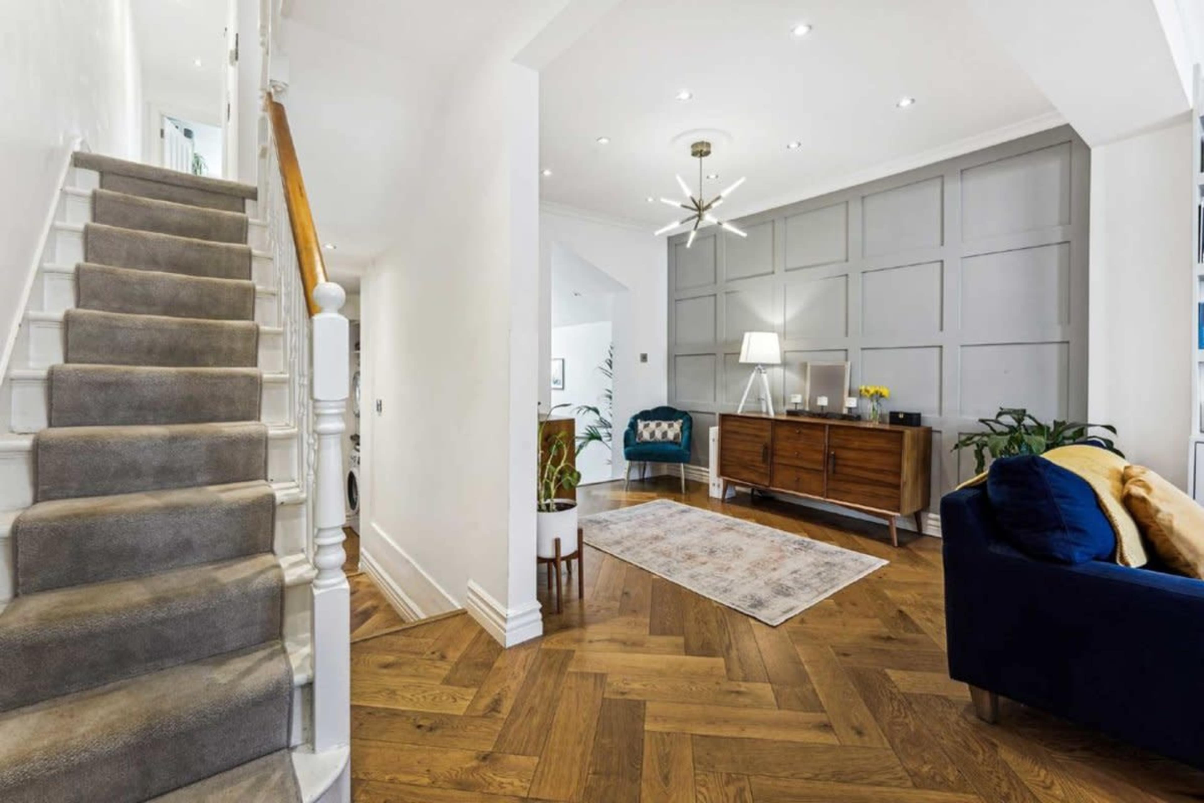 The image shows a well-lit foyer featuring a staircase, a wooden sideboard, and a blue couch, with plants and decorative elements in the space.