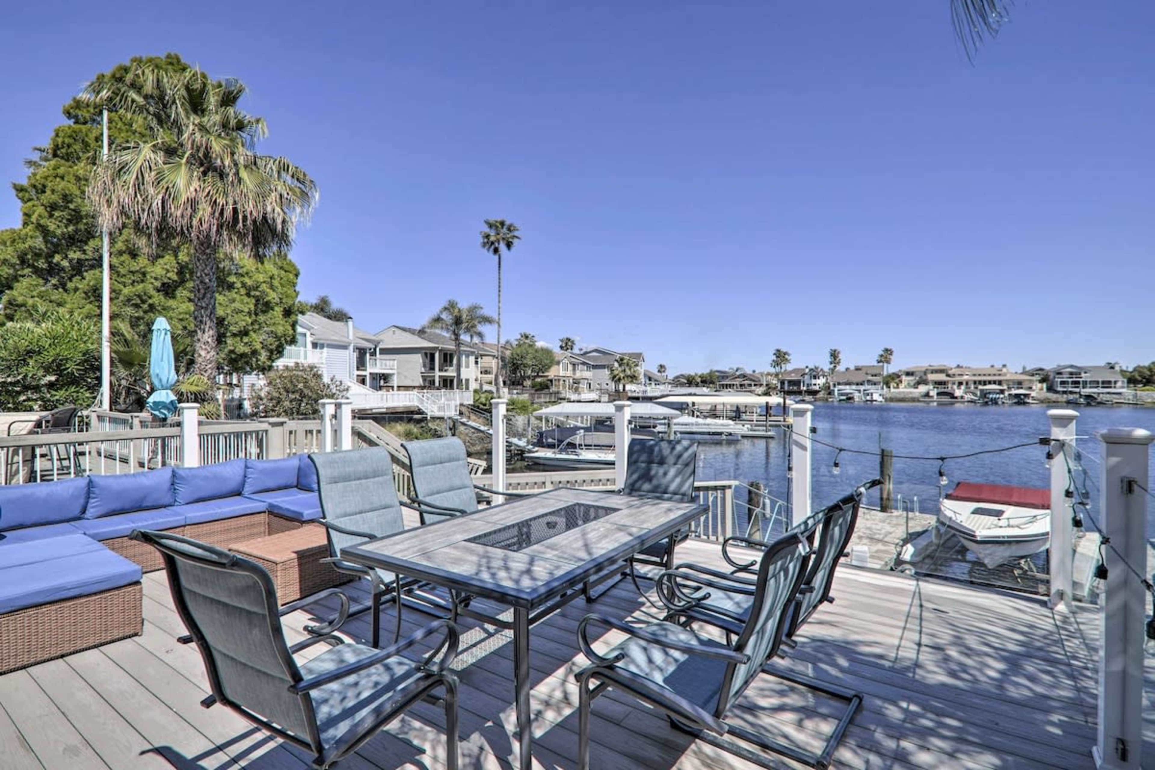 A patio equipped with a dining table and chairs overlooks a waterfront with boats and houses in the background.