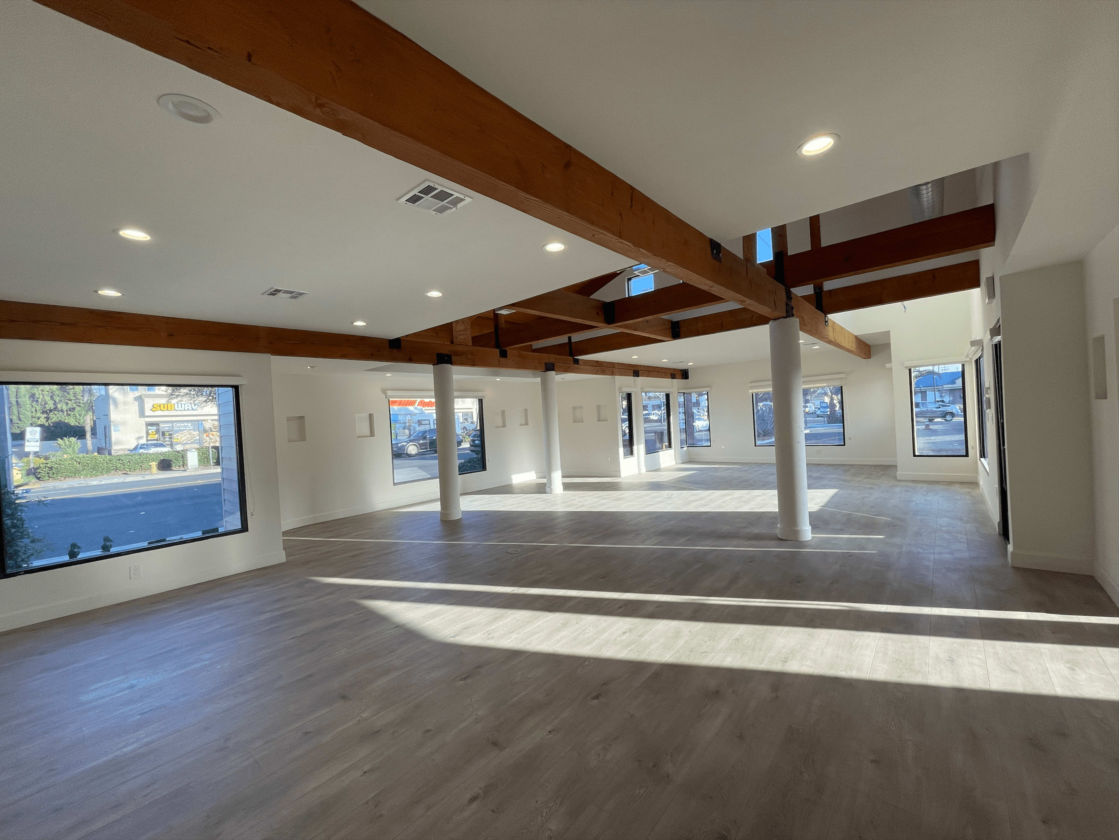The image shows an empty interior space with light wood beams and large windows allowing natural light to fill the room.