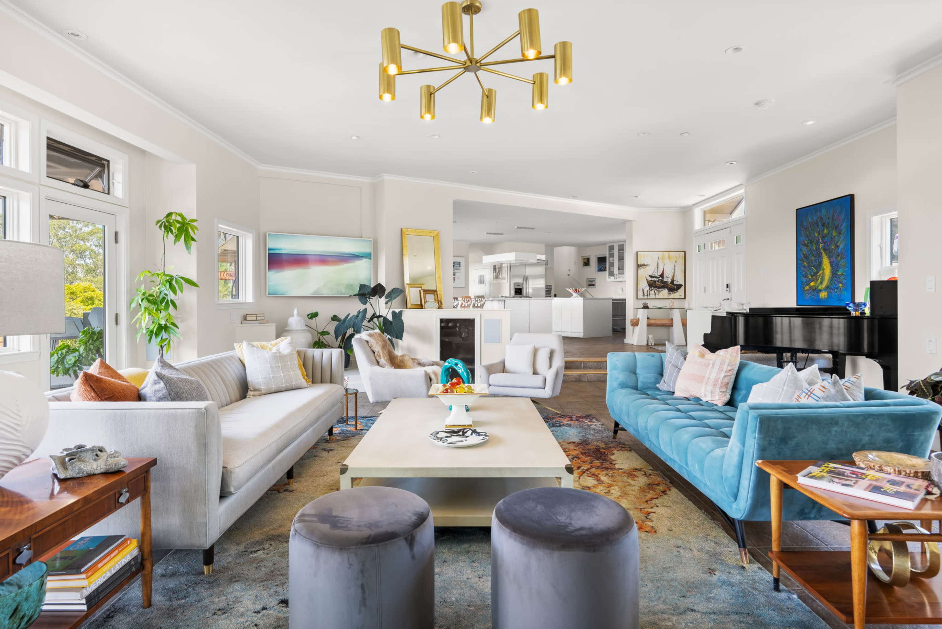 A bright living room featuring a mix of light-colored sofas, a blue velvet couch, a coffee table, and a piano, with large windows allowing natural light.