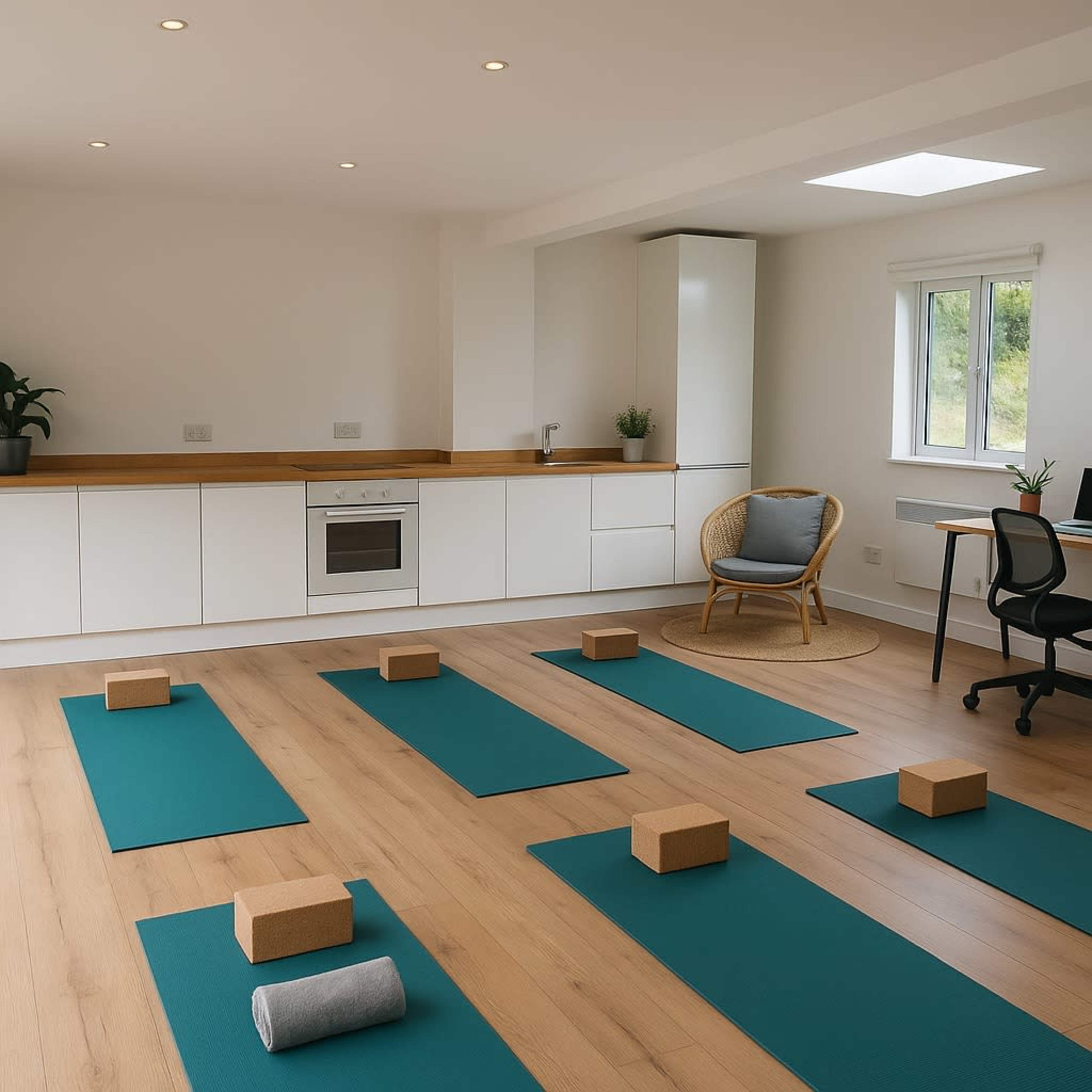 The image shows a spacious, bright yoga studio with multiple teal yoga mats arranged on a wooden floor, along with yoga blocks placed on each mat.