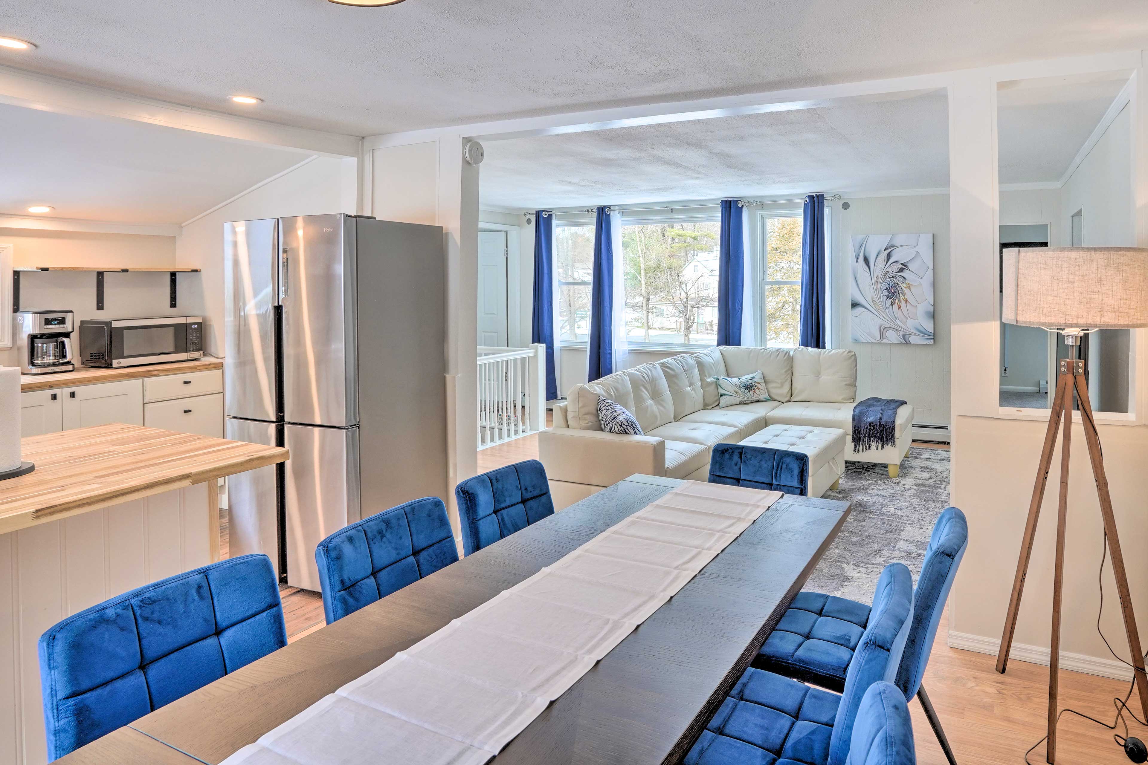 A modern open-concept living and dining area featuring a large dining table with blue chairs, a stainless steel refrigerator, a sectional sofa, and large windows allowing natural light.