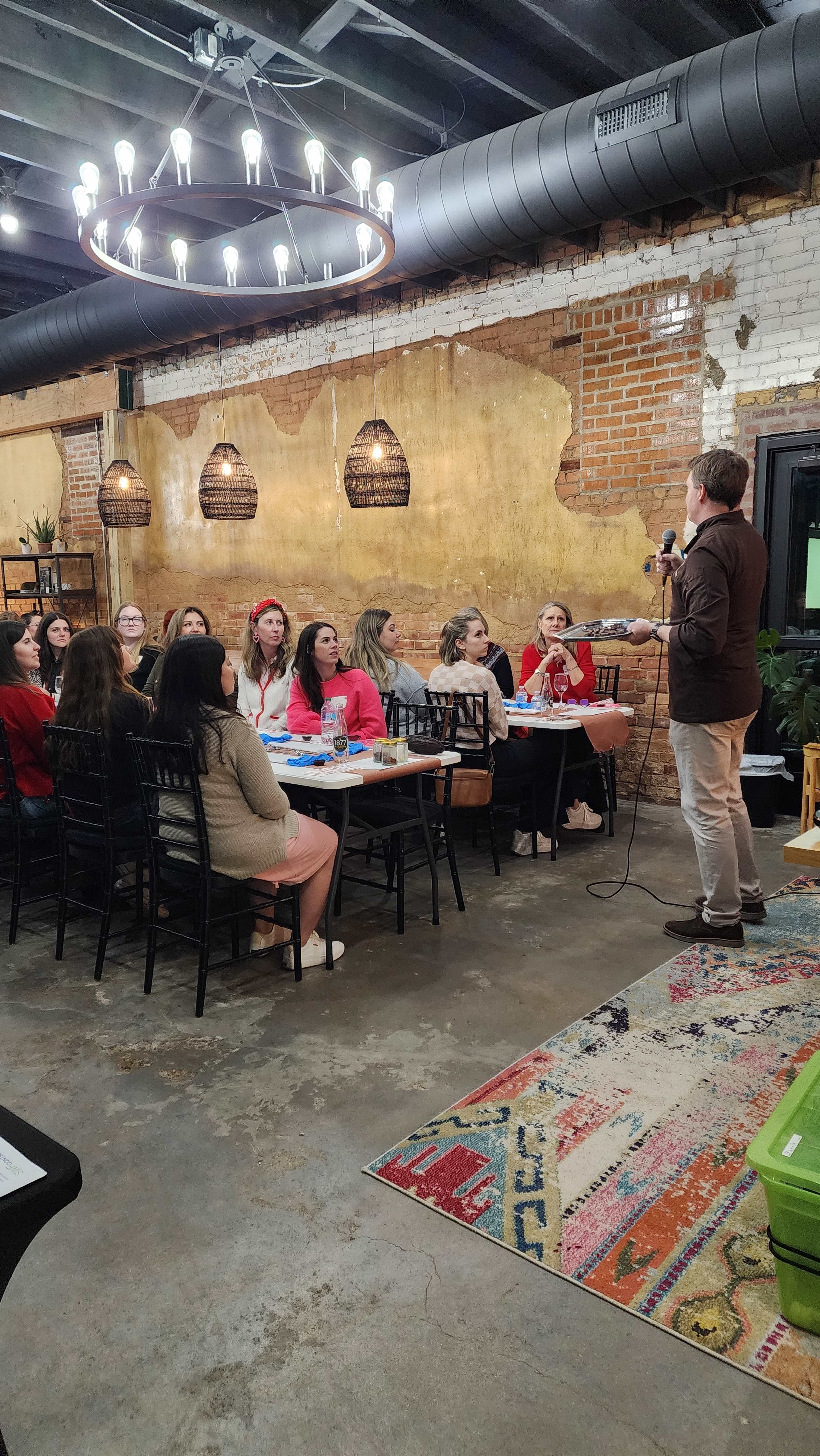 A man stands while speaking to a group of women seated at tables in a rustic interior with exposed brick and pendant lighting.