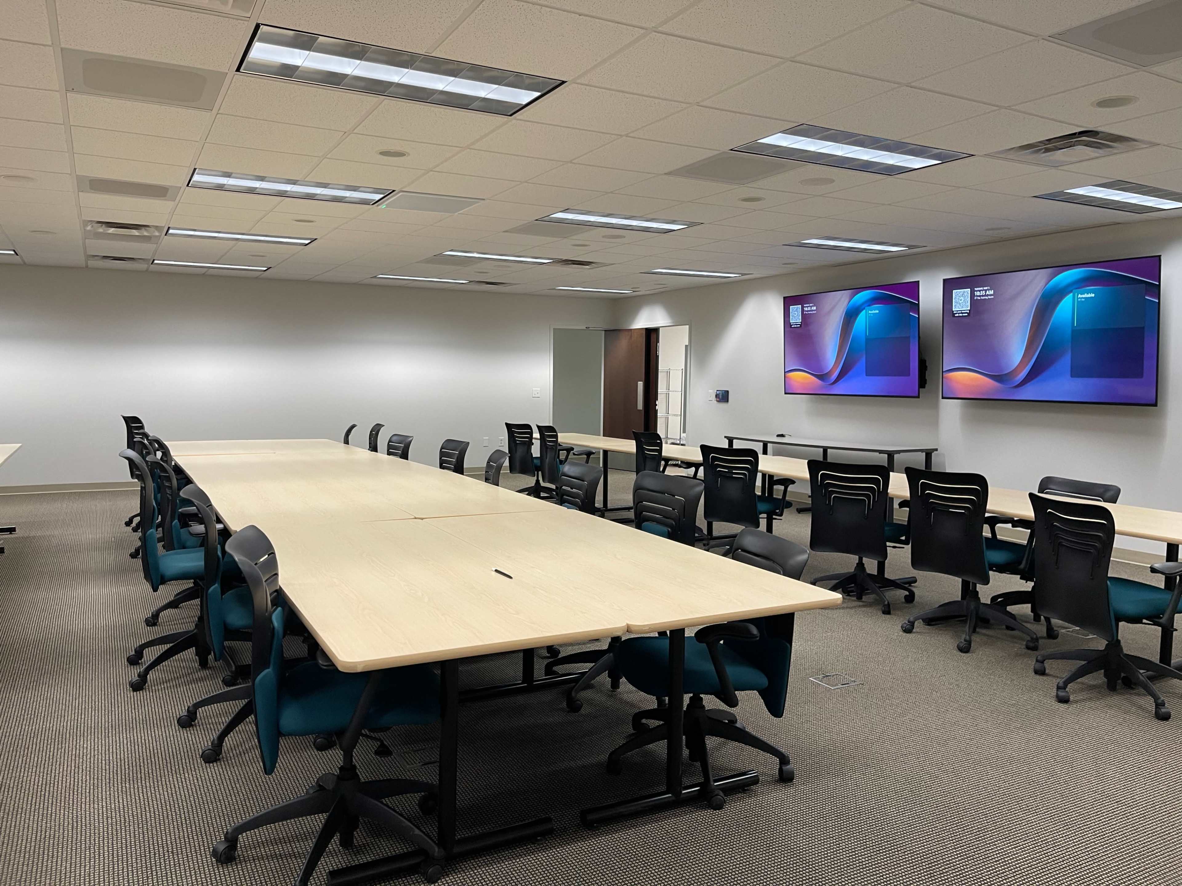 The image shows a large, empty conference room with several rows of tables and chairs, along with two display screens mounted on the wall.
