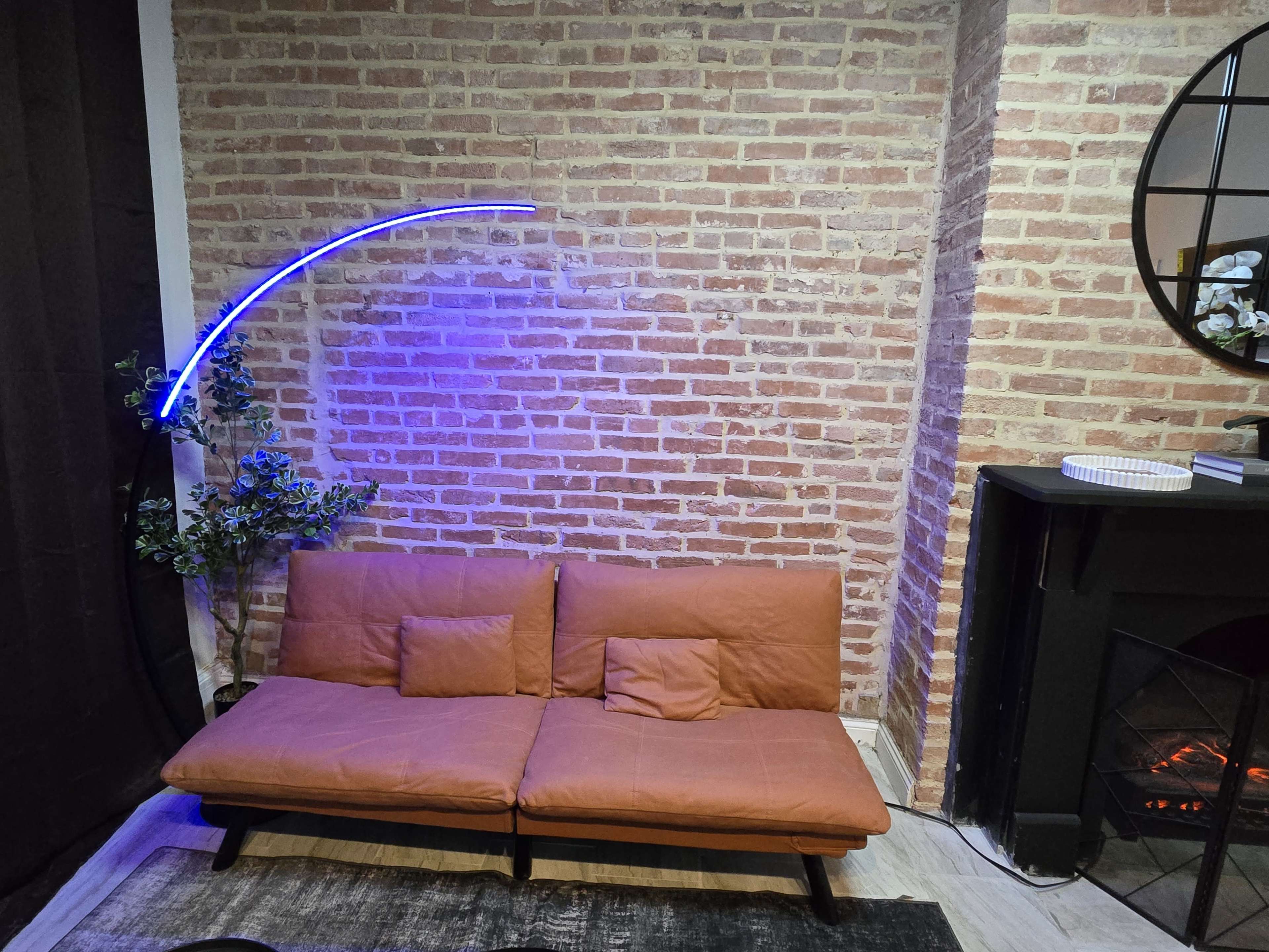 The image shows a modern living space with a brown sofa, a curved blue neon light, a potted plant, and a fireplace against a brick wall.