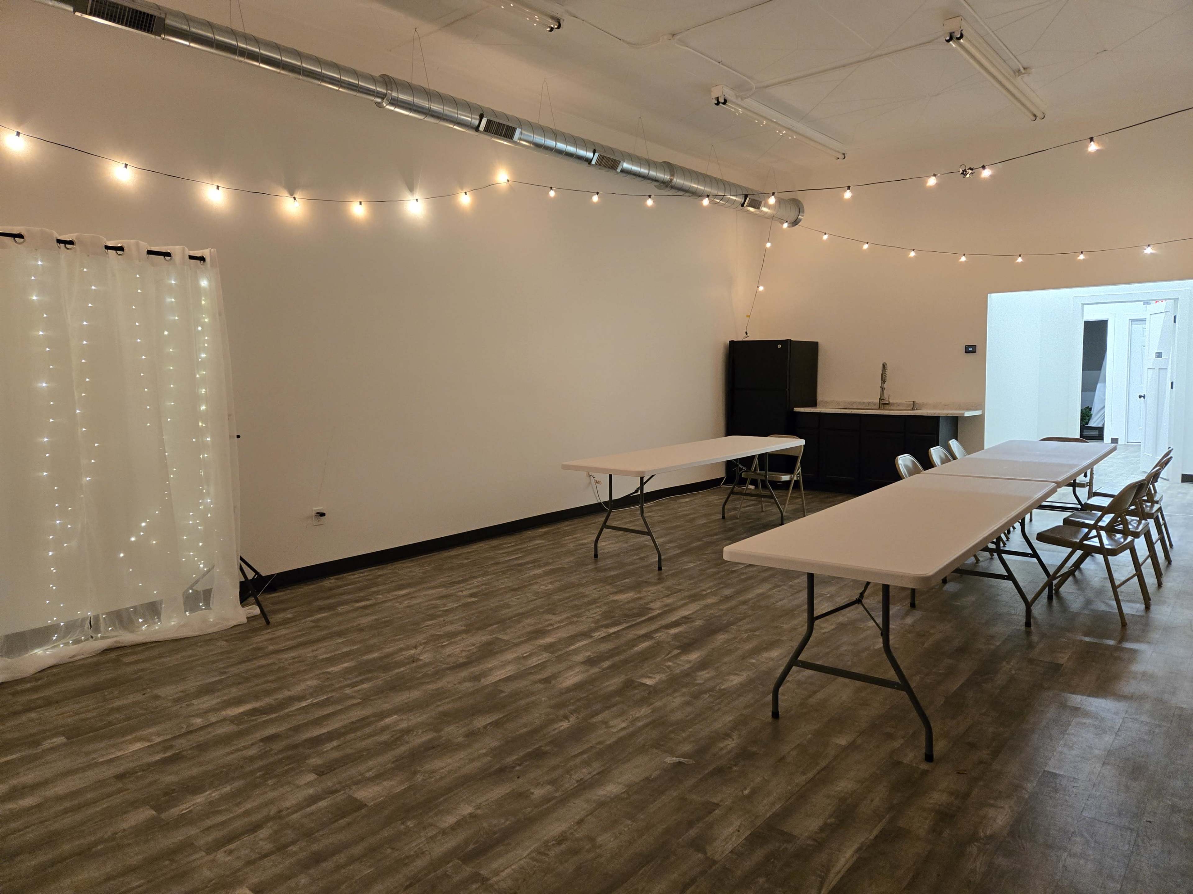 The image shows a spacious indoor area with several rectangular tables set up, string lights hanging from the ceiling, and a small kitchenette visible in the background.