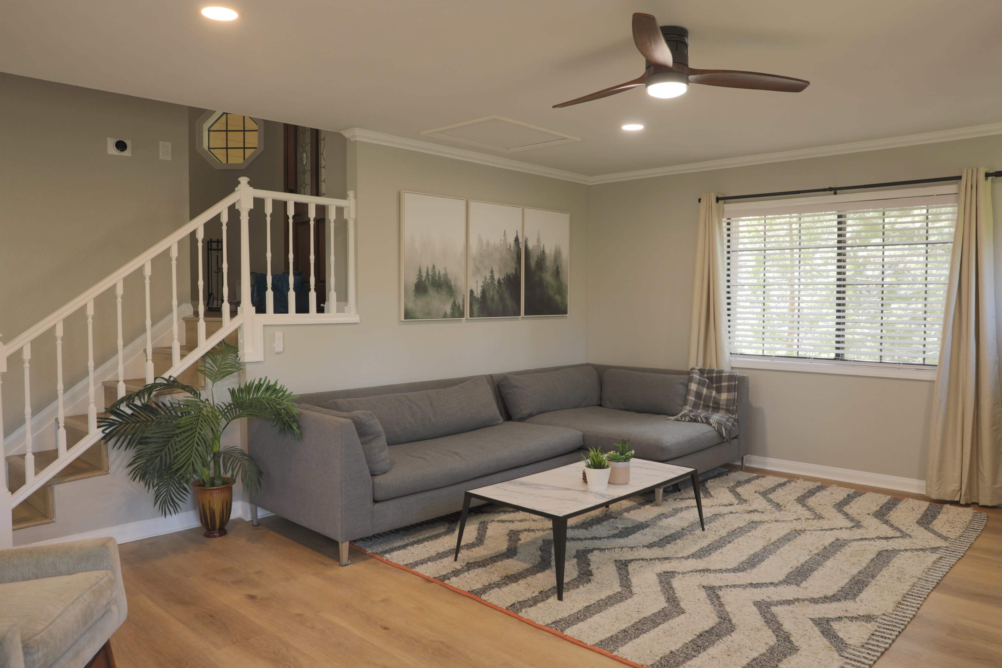 A modern living room features a gray sectional sofa, a coffee table on a patterned rug, and large windows with curtains.