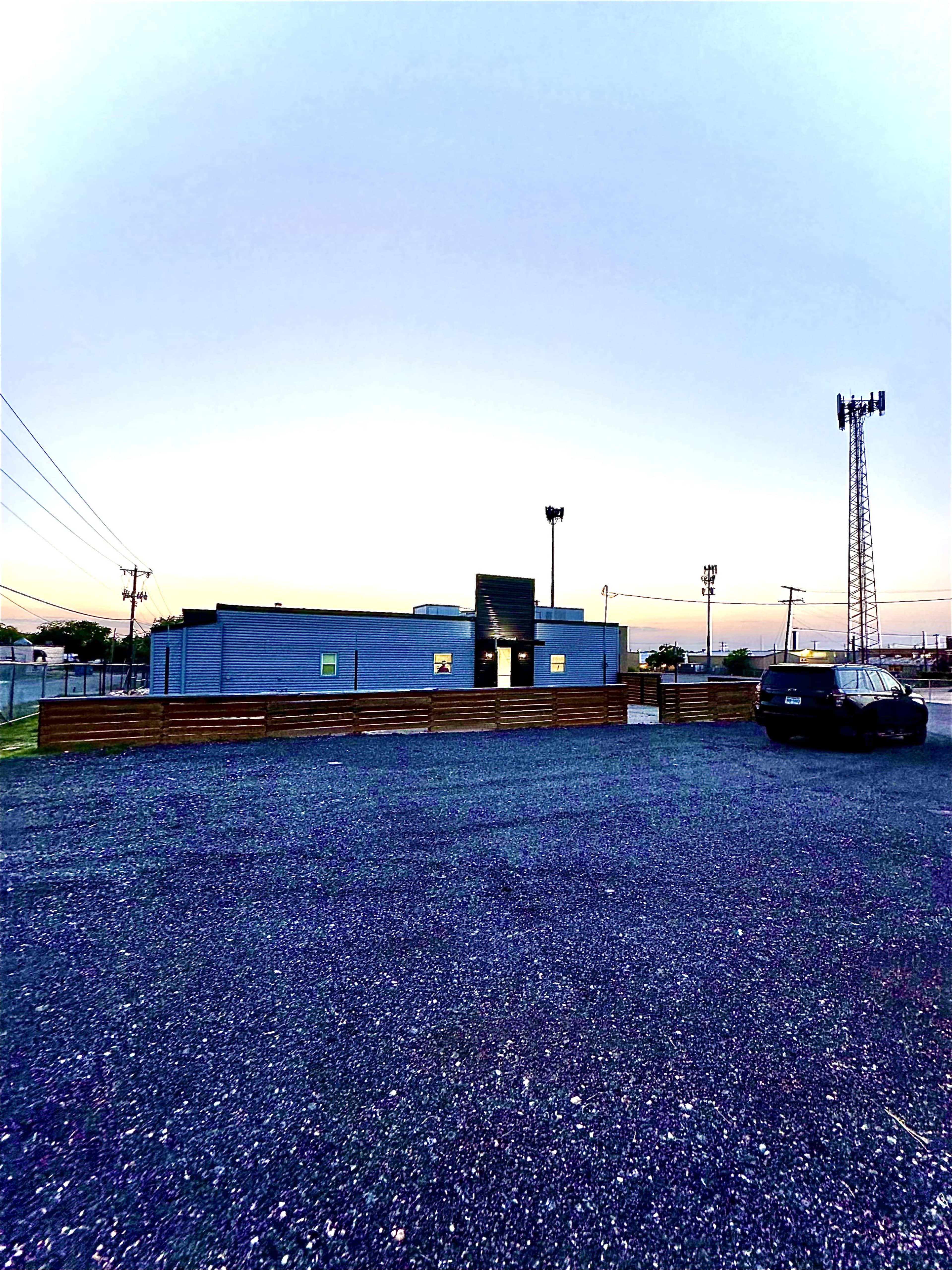 A gray building surrounded by a gravel parking lot, with a cell tower in the background and a clear sky at dusk.