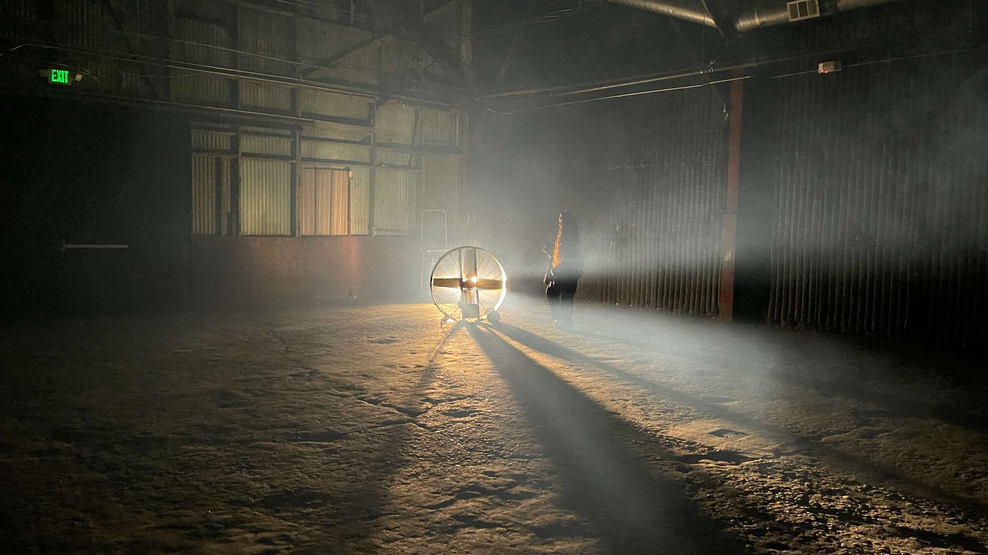 A figure stands near a circular light source in a dimly lit industrial space, casting long shadows on the ground.