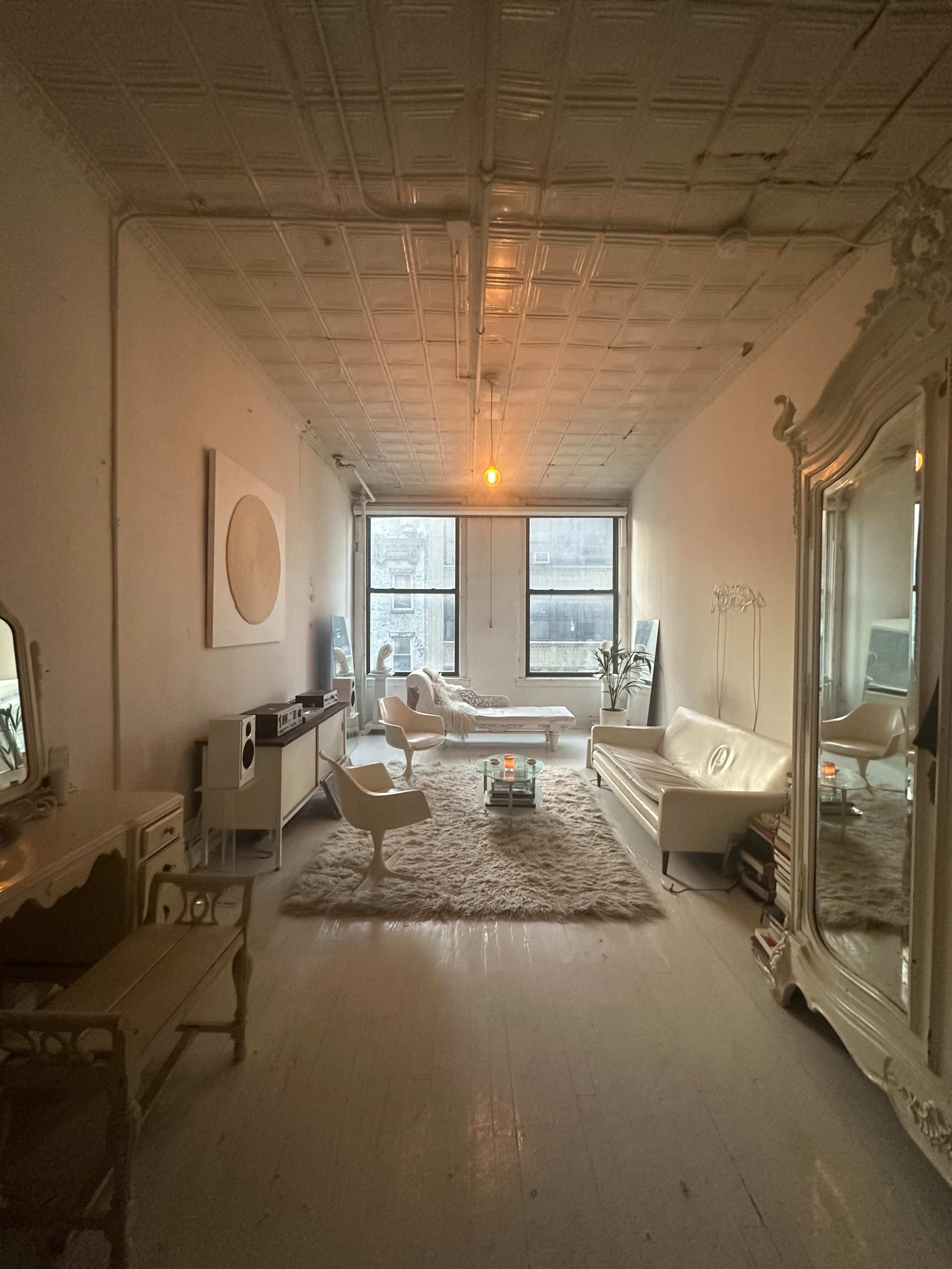 The image shows a spacious, well-lit room with large windows, featuring a minimalist design, a white sofa, a glass coffee table, and a large mirror against one wall.