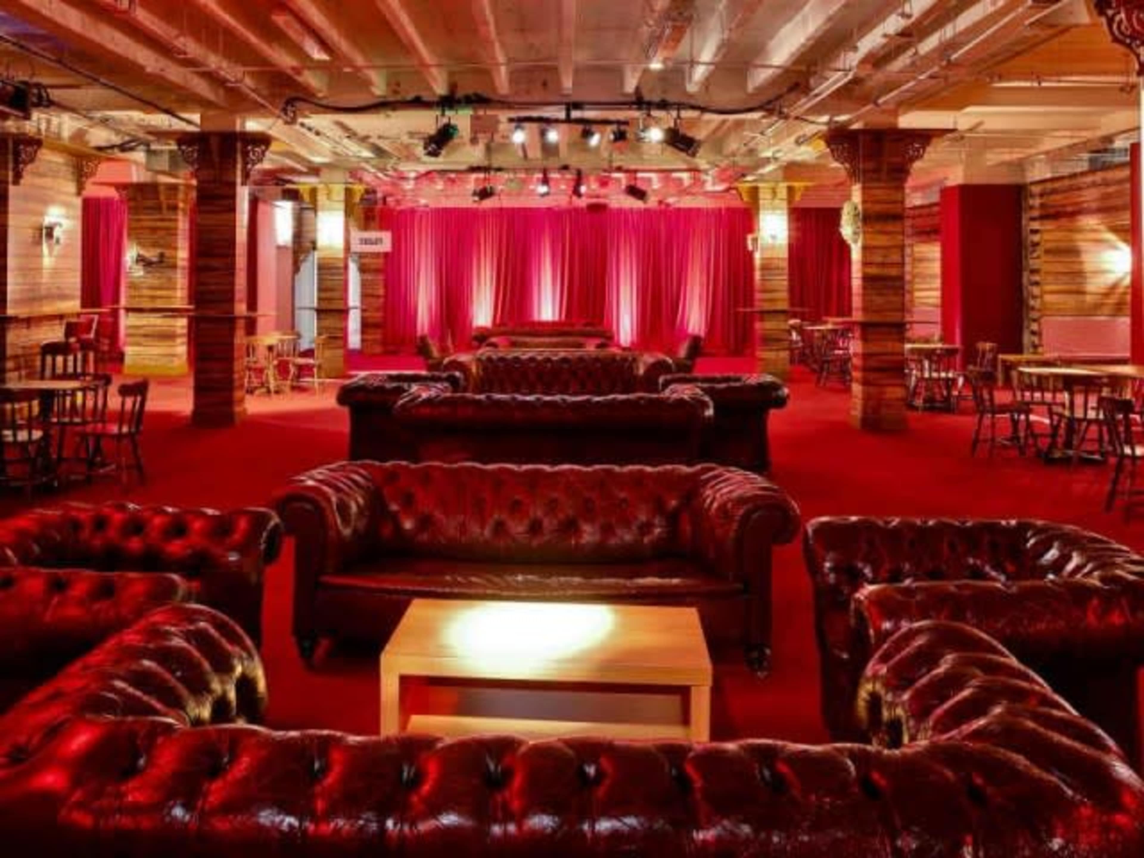 The interior of a lounge features red velvet seating arranged around a central table, with a stage and curtains at the far end.
