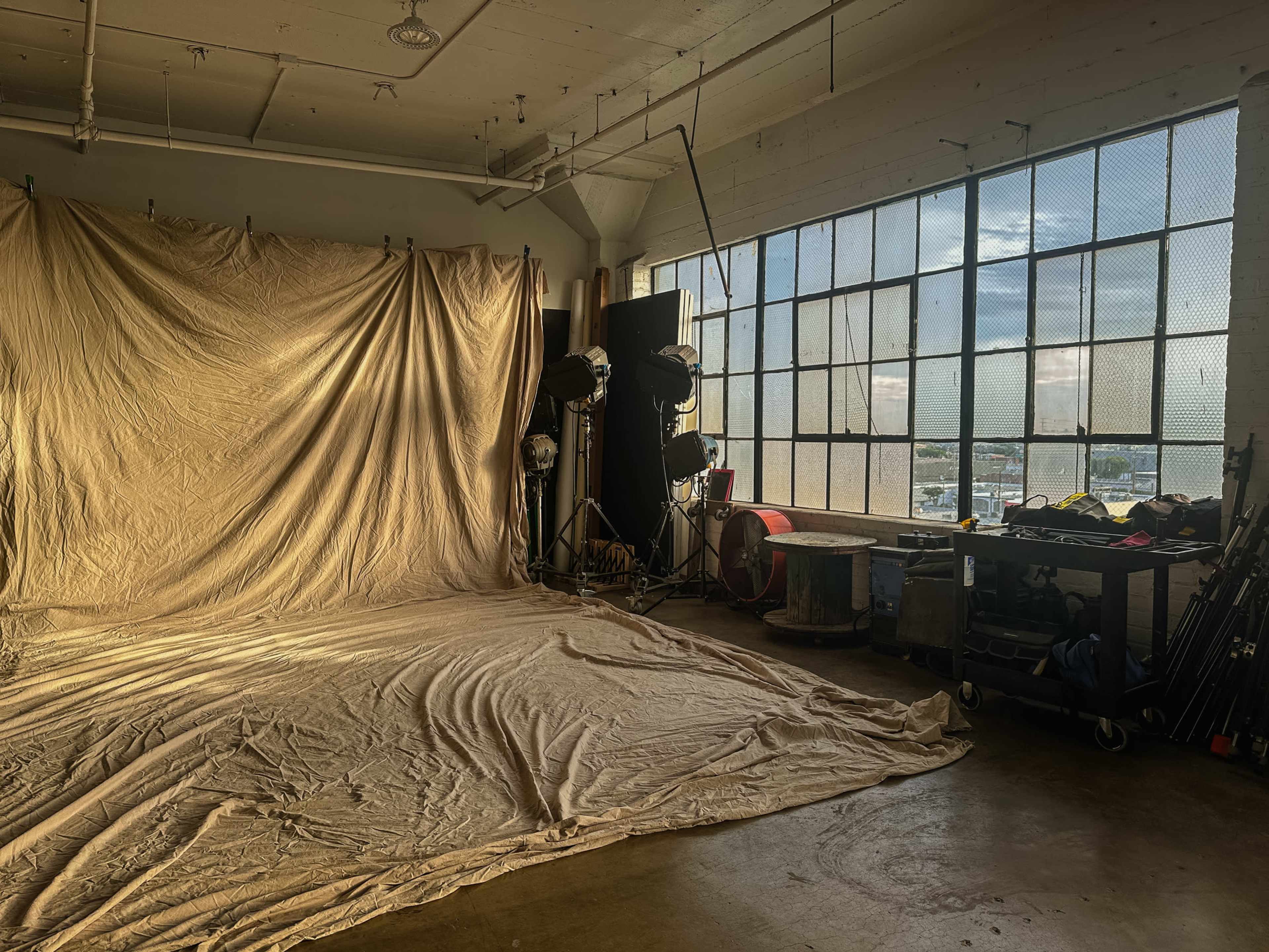 The image shows a photography studio with a wrinkled fabric backdrop, studio lights, and equipment near a large window letting in natural light.