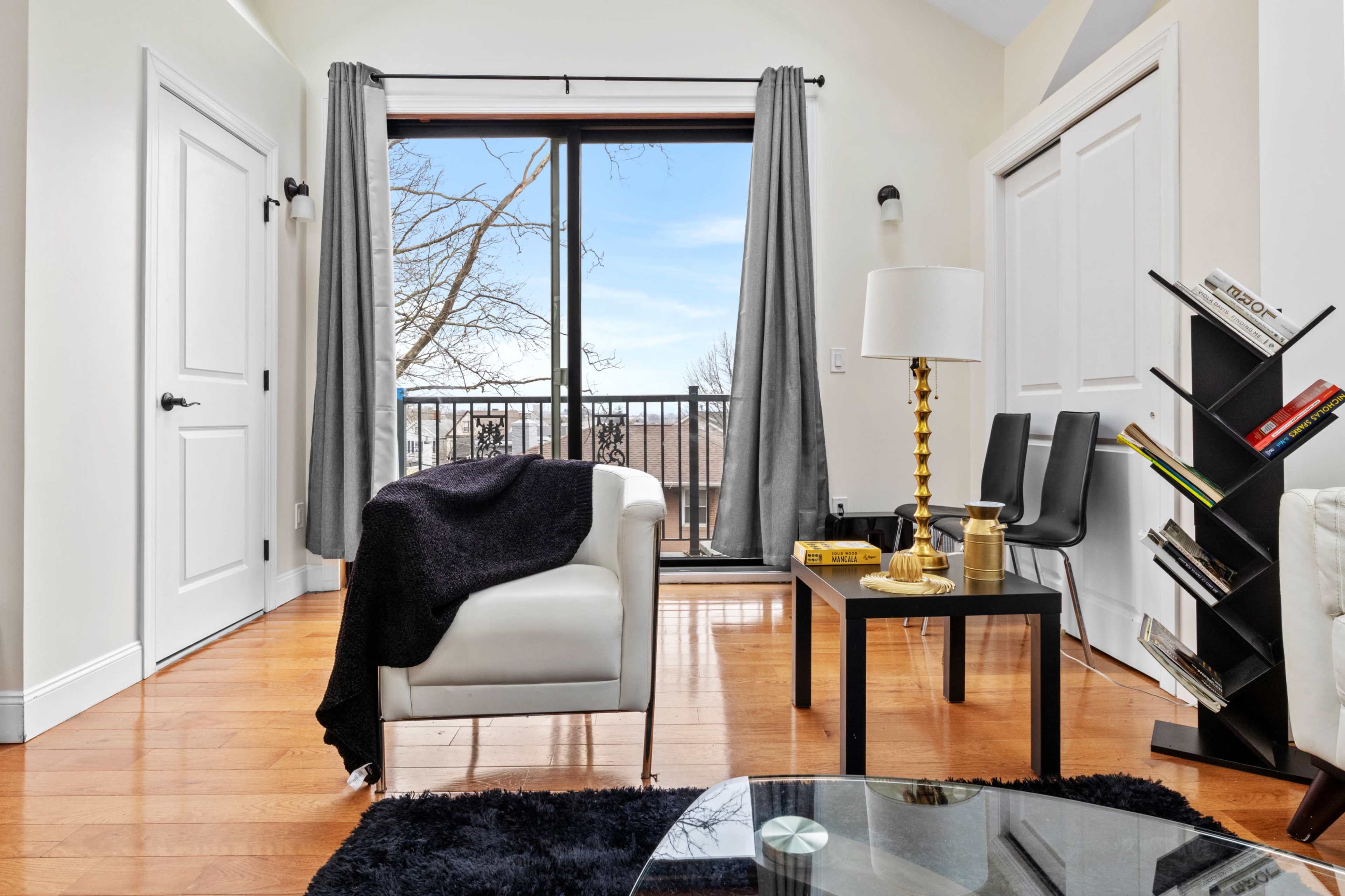 A bright living room features a white armchair with a blanket, a black side table, and a floor-to-ceiling window with gray drapes overlooking a tree-lined view.