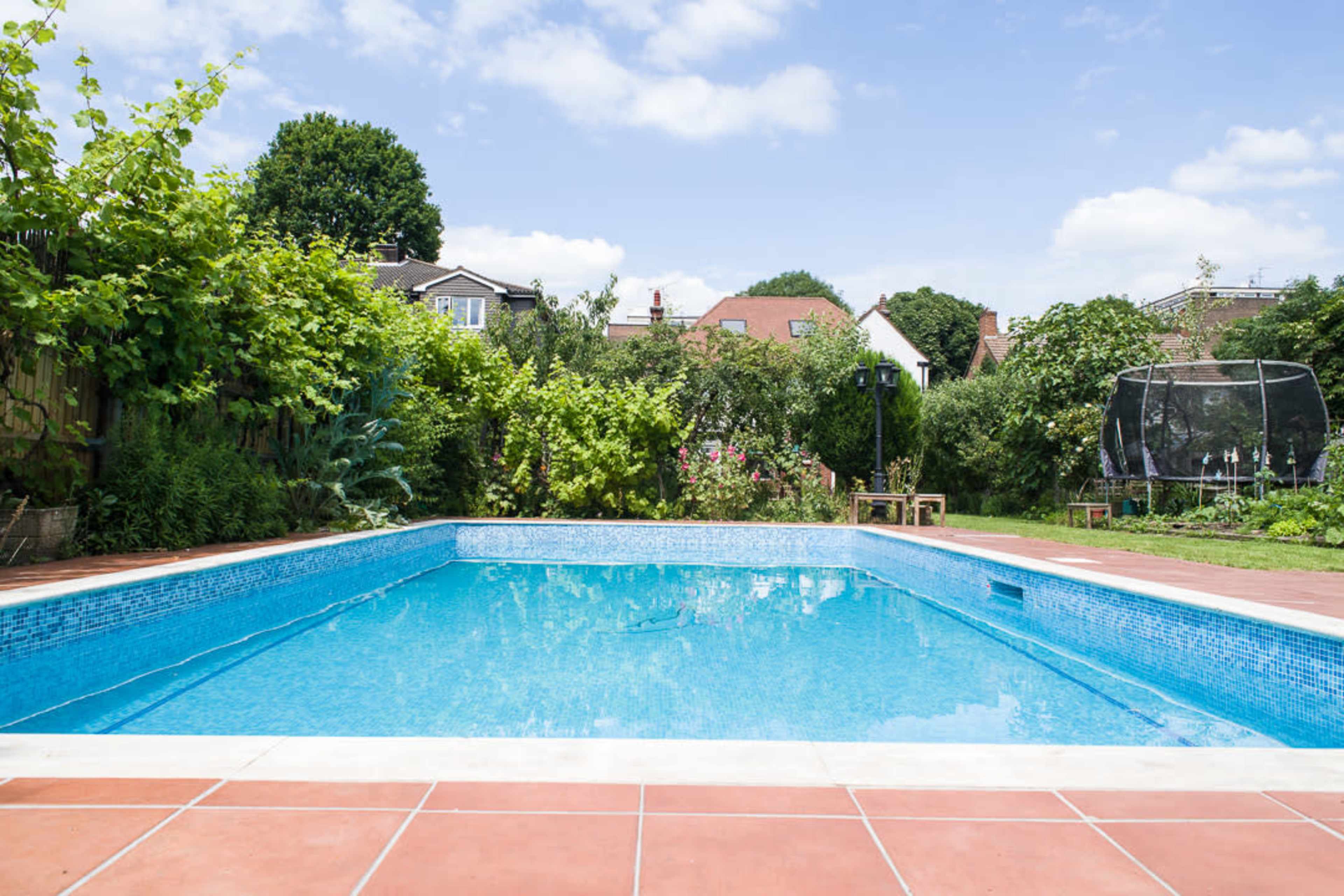 A clear blue swimming pool surrounded by lush greenery and residential houses in the background.