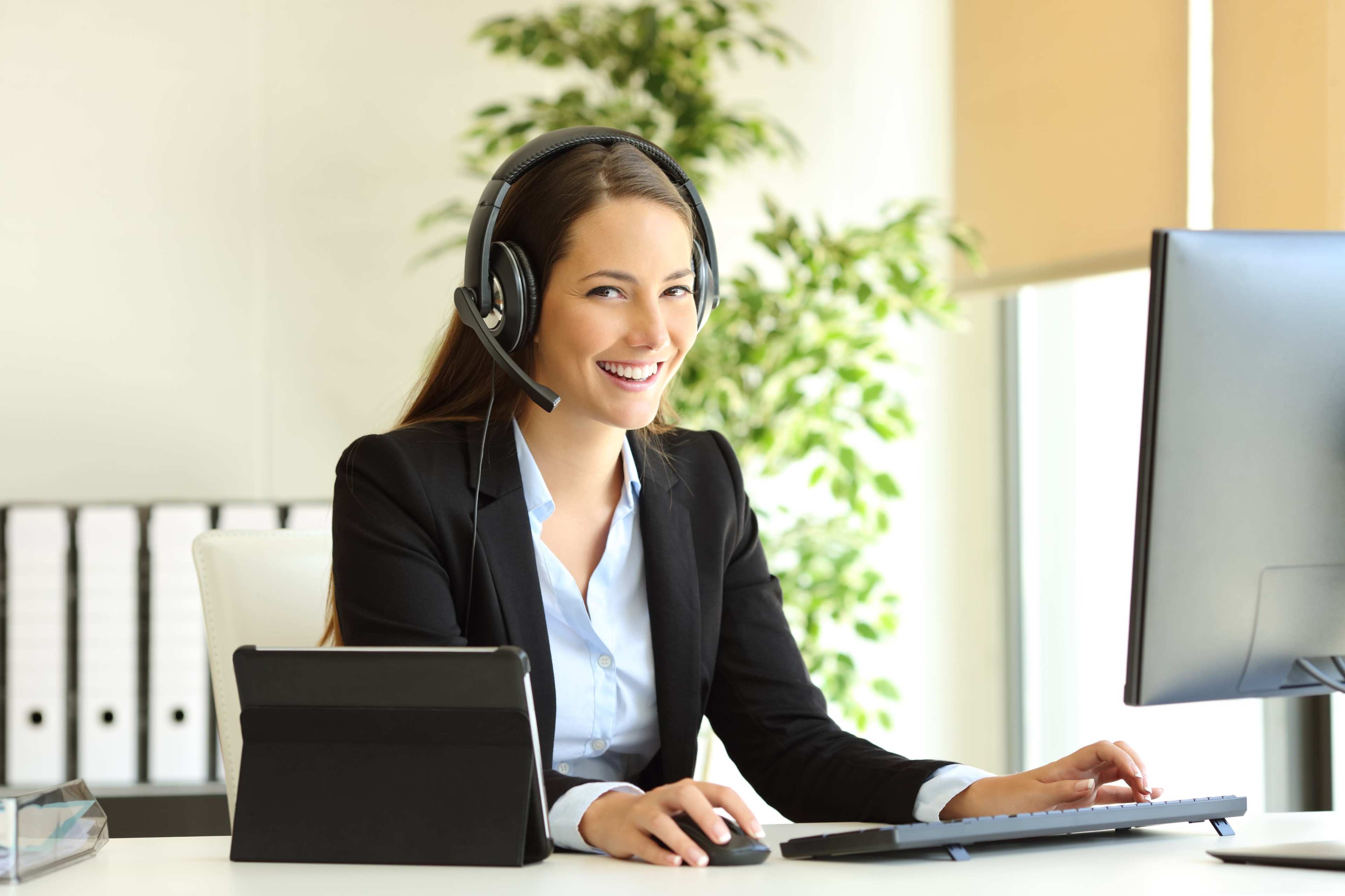 A woman wearing a headset sits at a desk with a computer and tablet, smiling as she works in a bright office environment.