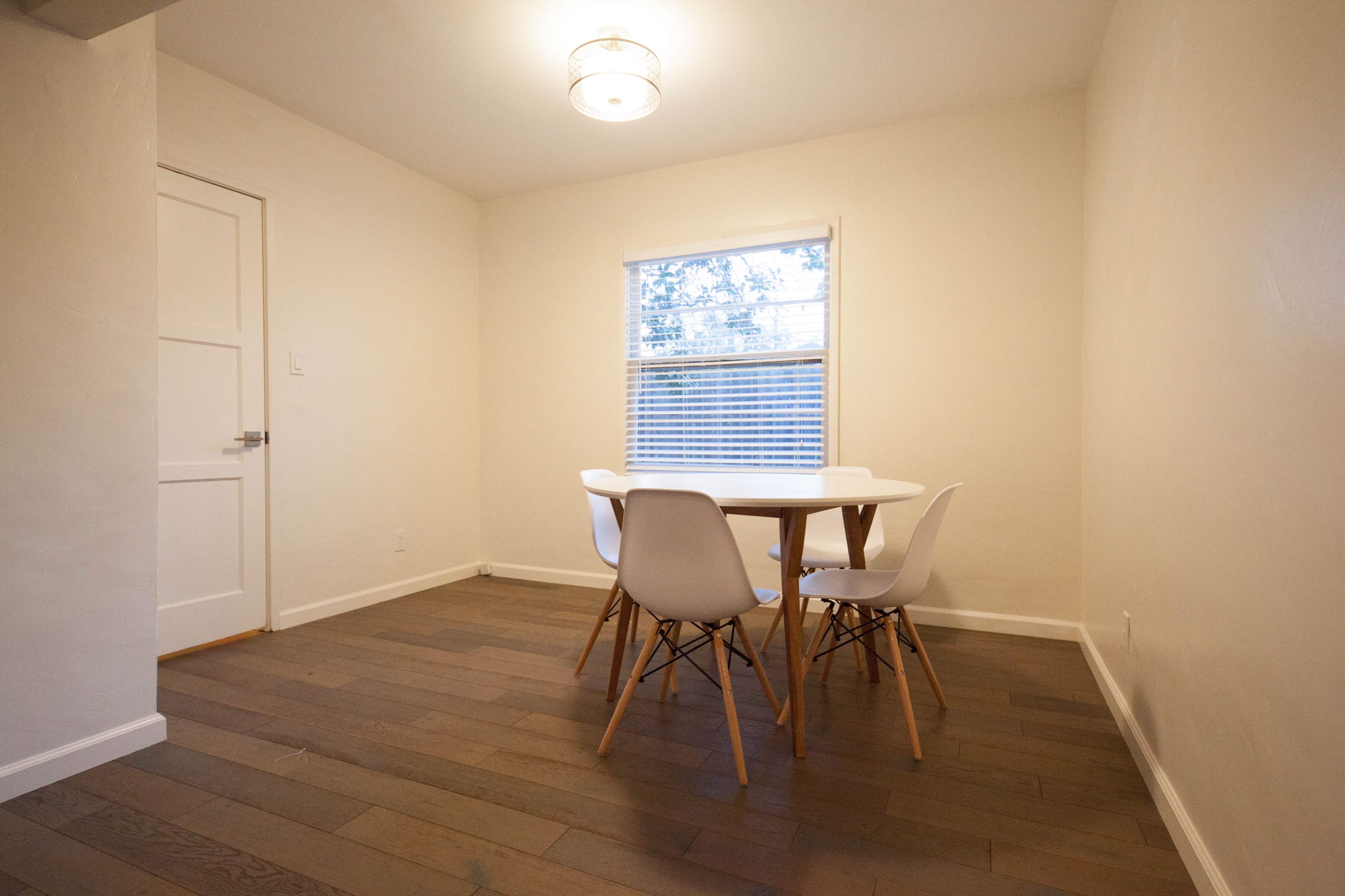 A simple dining area with a round table and four white chairs, set against a wall with a window that lets in natural light.