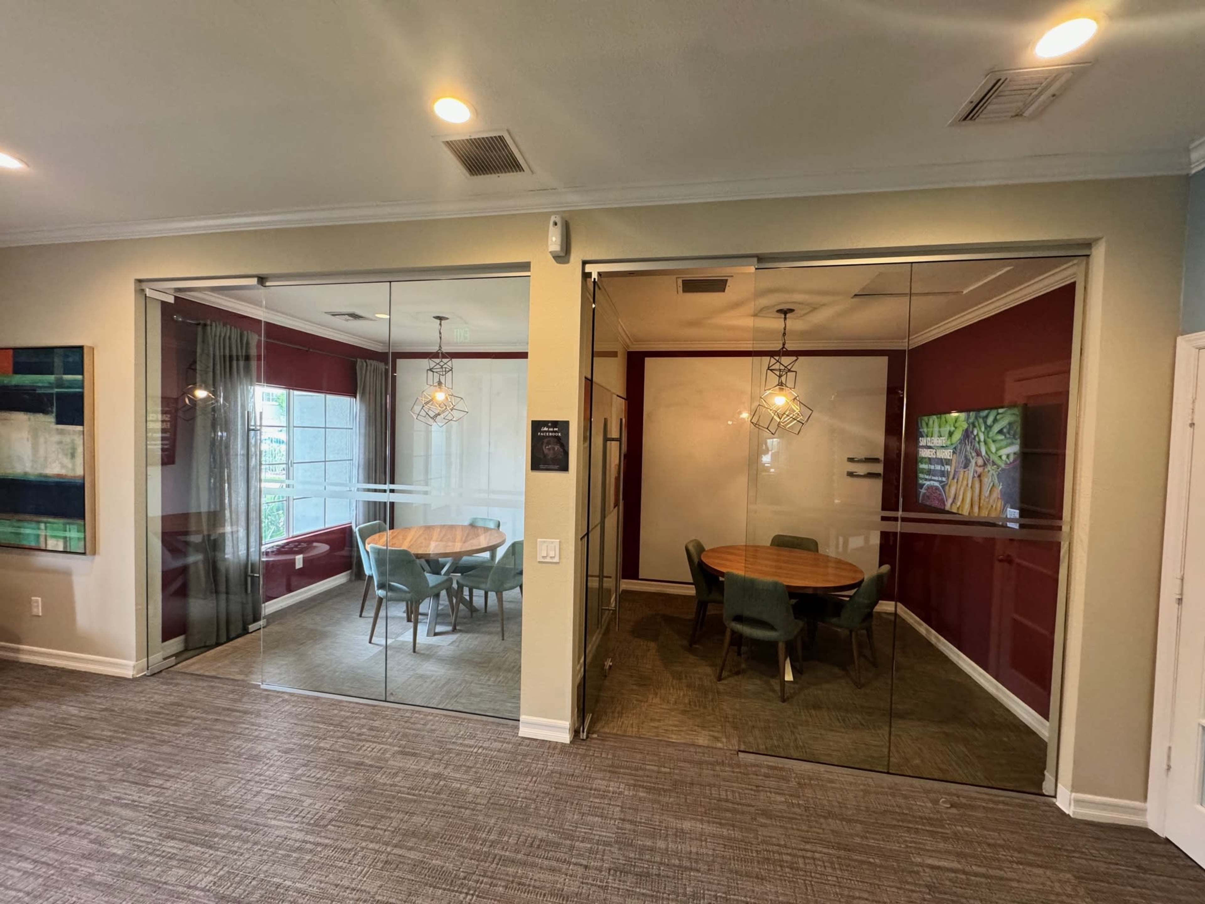 A modern office space with two glass-walled conference rooms, one featuring a round wooden table and green chairs, and the other a seating area with a view outside.